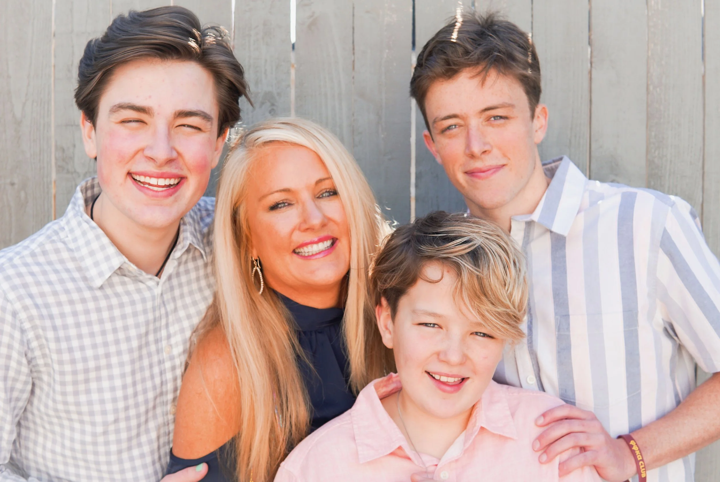 Group of four smiling people, including a woman and three teenage boys, standing outdoors in front of a wooden fence.