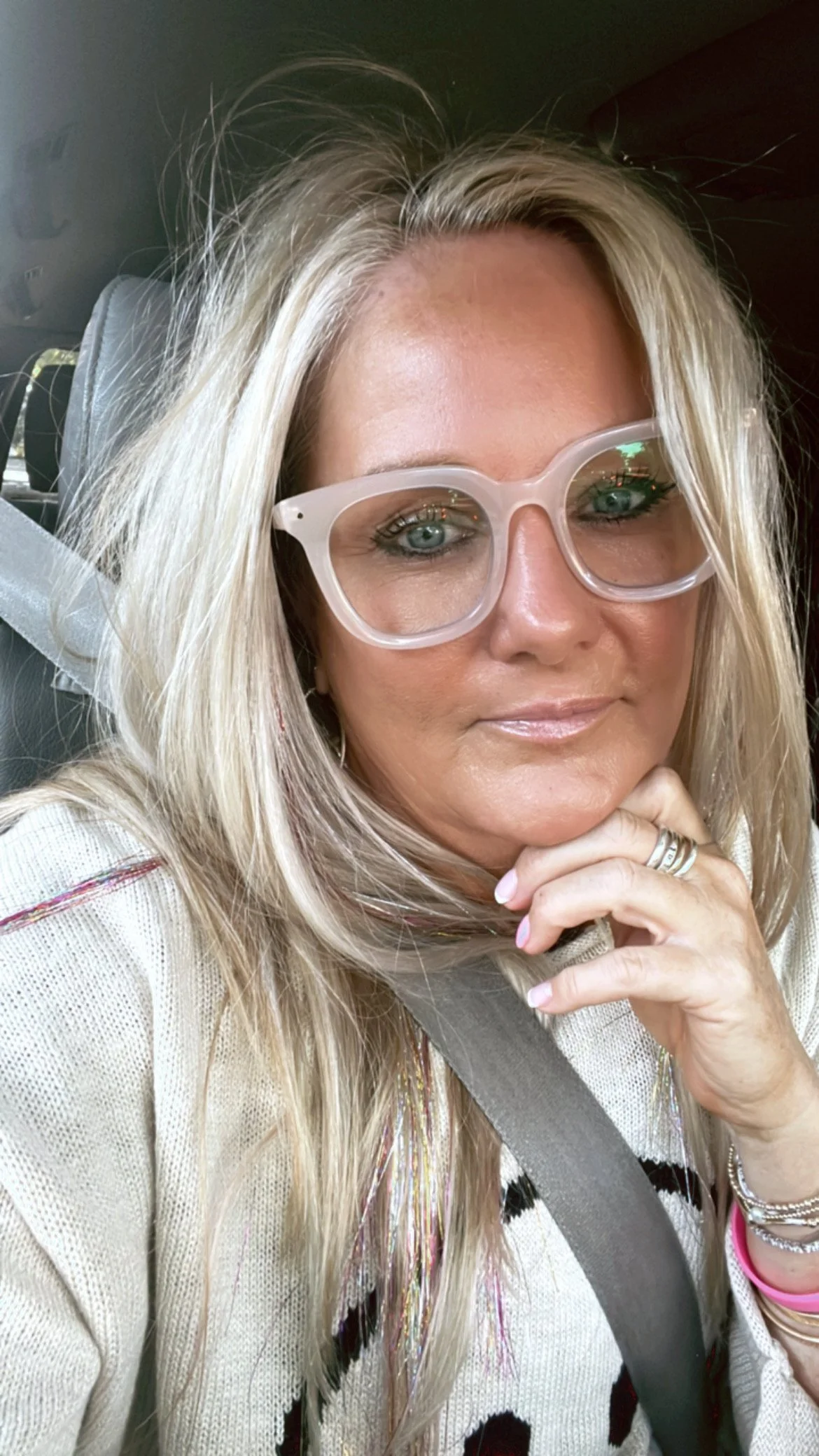 A woman with long blonde hair wearing large clear glasses, sitting in a car and resting her chin on her hand, wearing rings and bracelets.