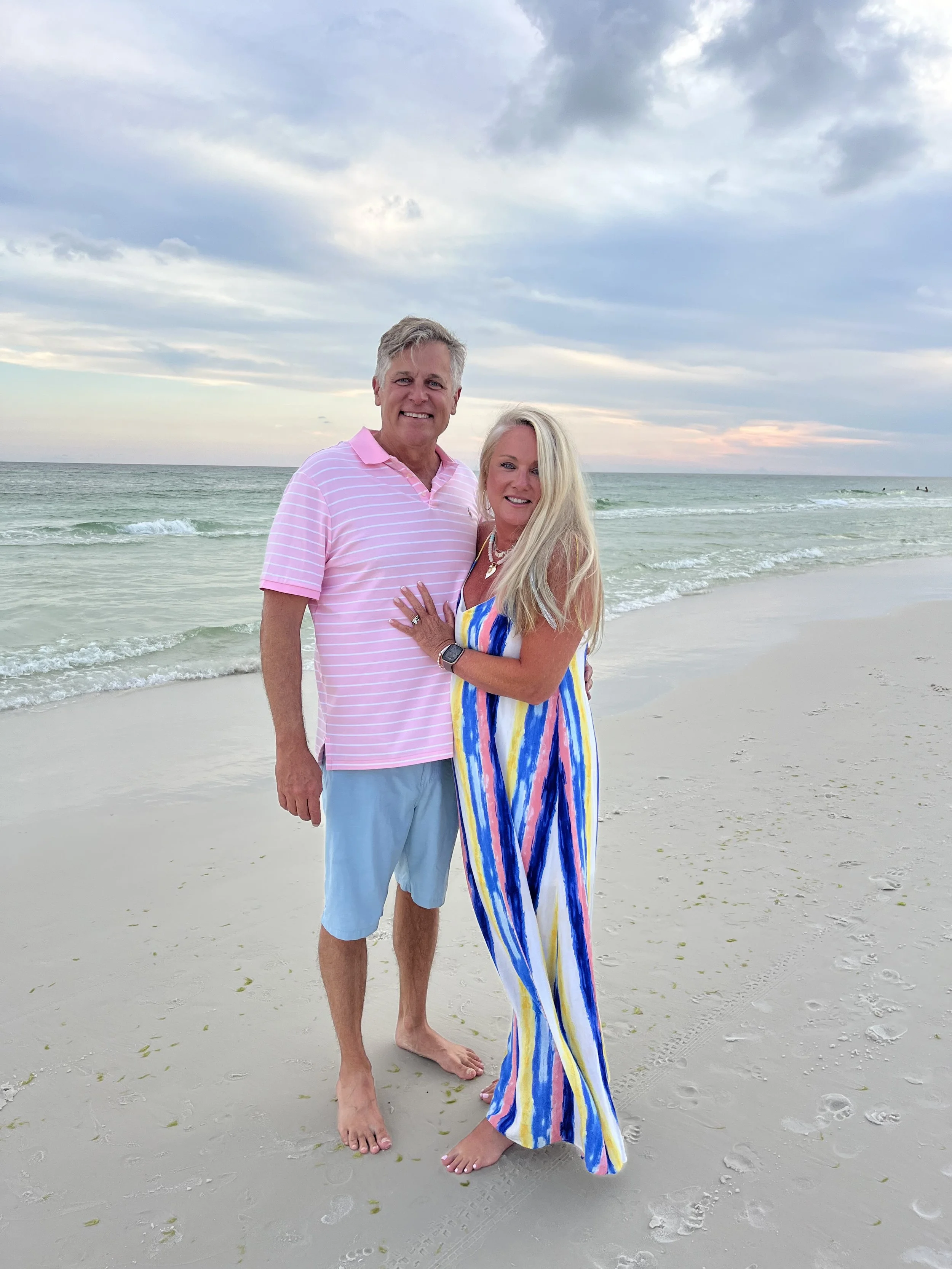 A smiling middle-aged man and woman standing on a beach with the ocean and cloudy sky in the background. The man is wearing a pink striped polo shirt and light blue shorts, and the woman is wearing a colorful striped dress. They are barefoot and stan