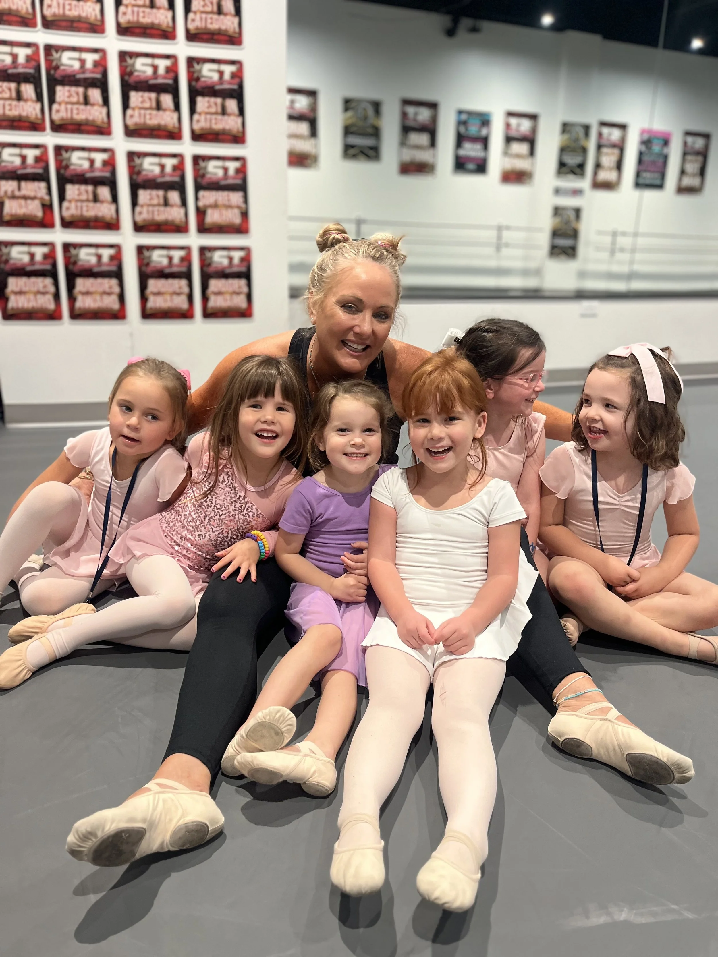Six young girls and an adult woman sitting on the floor of a dance studio, smiling and posing for the photo. The studio has posters on the wall and mirrored walls.