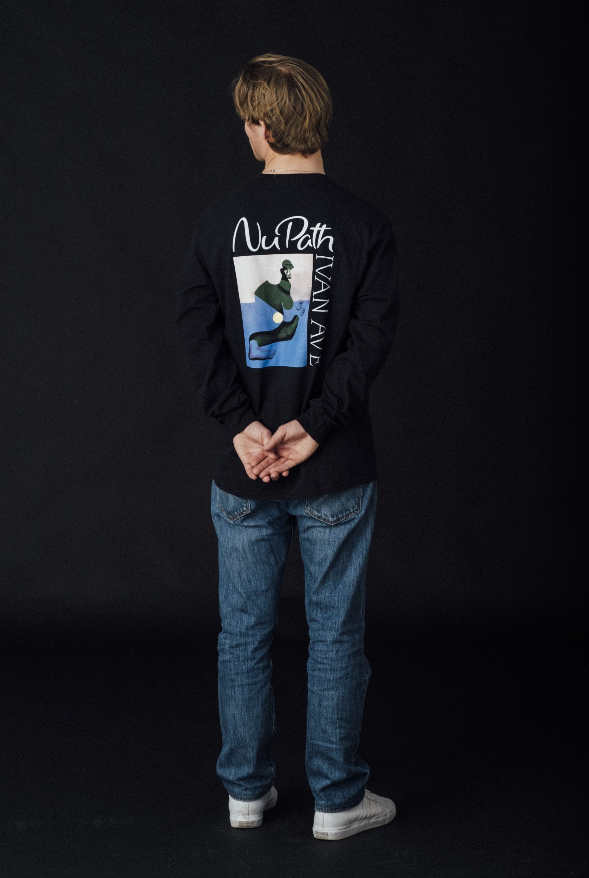 A person standing with back facing the camera against a black background, wearing a black long-sleeve shirt with colorful graphic and text, blue jeans, and white sneakers.