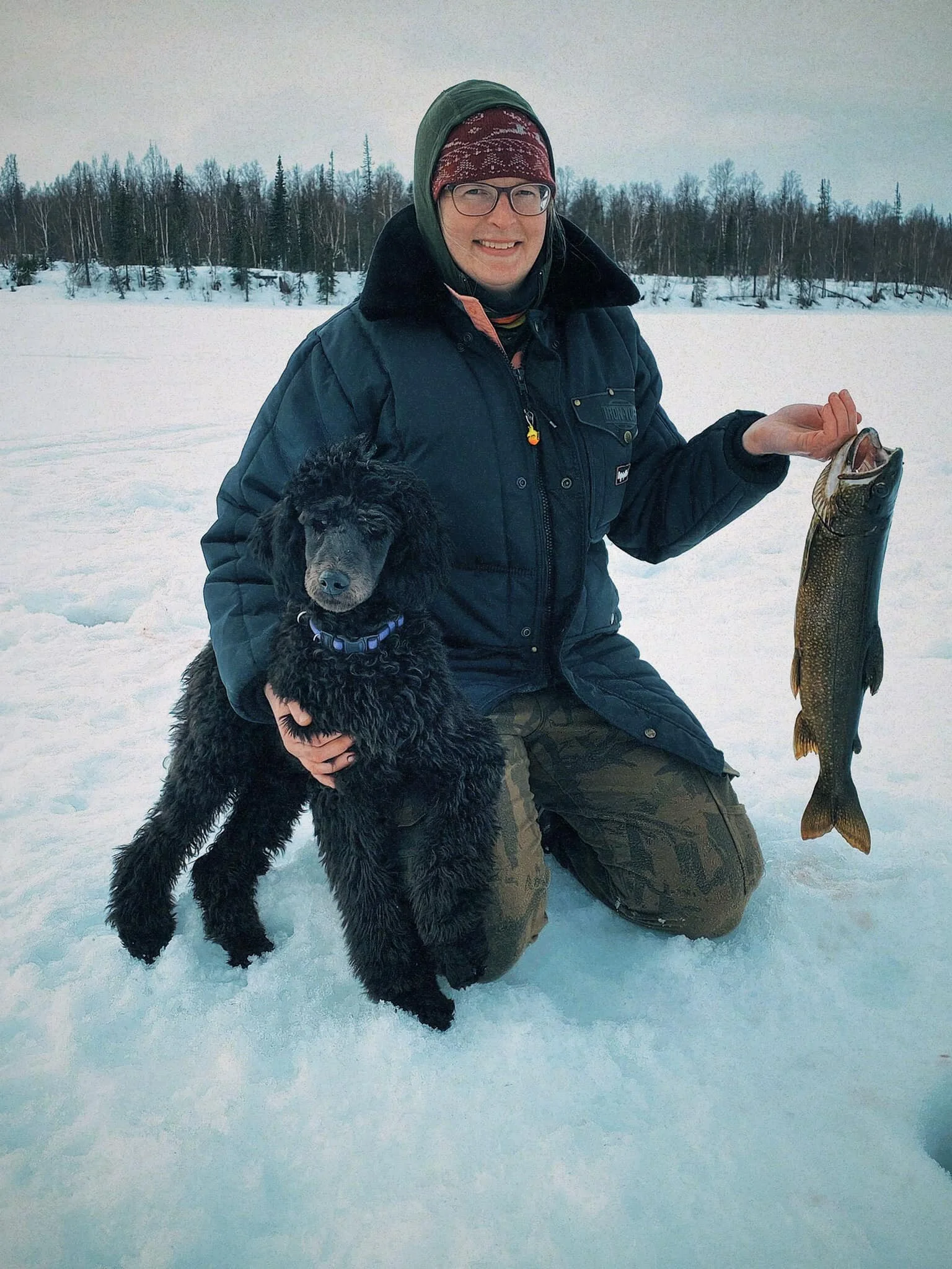 A woman ice fishing is holding a fish while hugging her black standard poodle puppy. The poodle and the woman are kneeling on a lake that has been iced over.