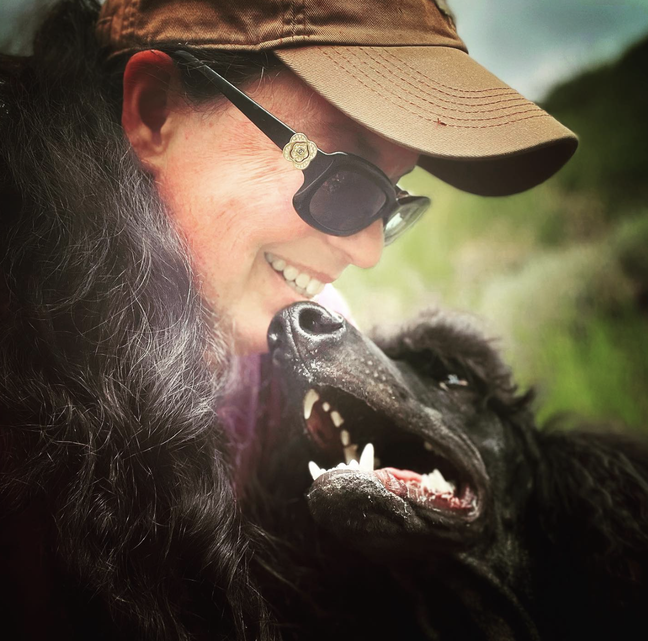 A woman wearing sunglasses and a baseball cap shares a joyful moment with her black Standard Poodle, smiling as her dog looks up at her with an open-mouthed grin.