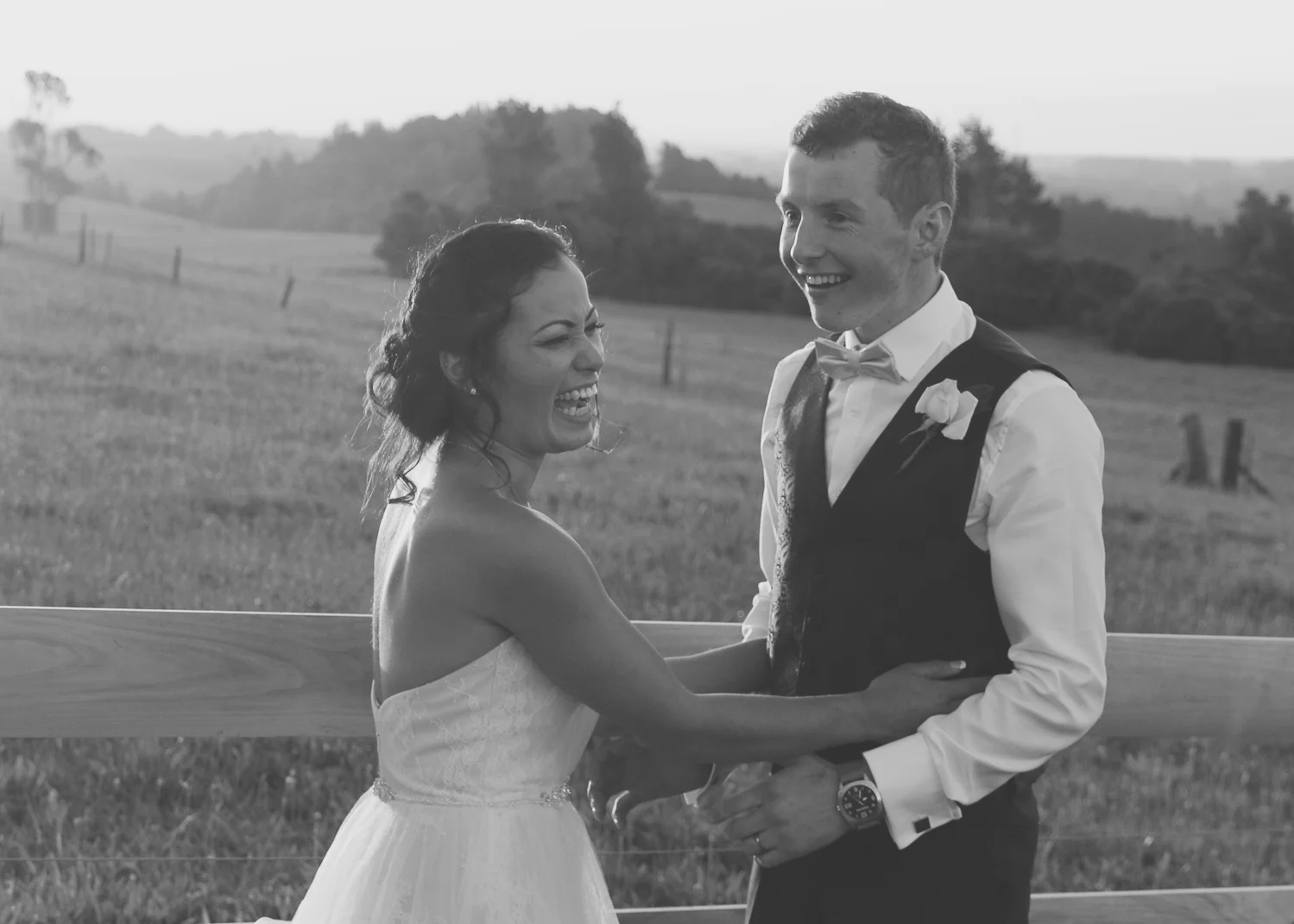 bride_and_groom_on_wedding_day_at_sunset_laughing_and_in_love_by_fence_black_and_white_on_farm.jpg