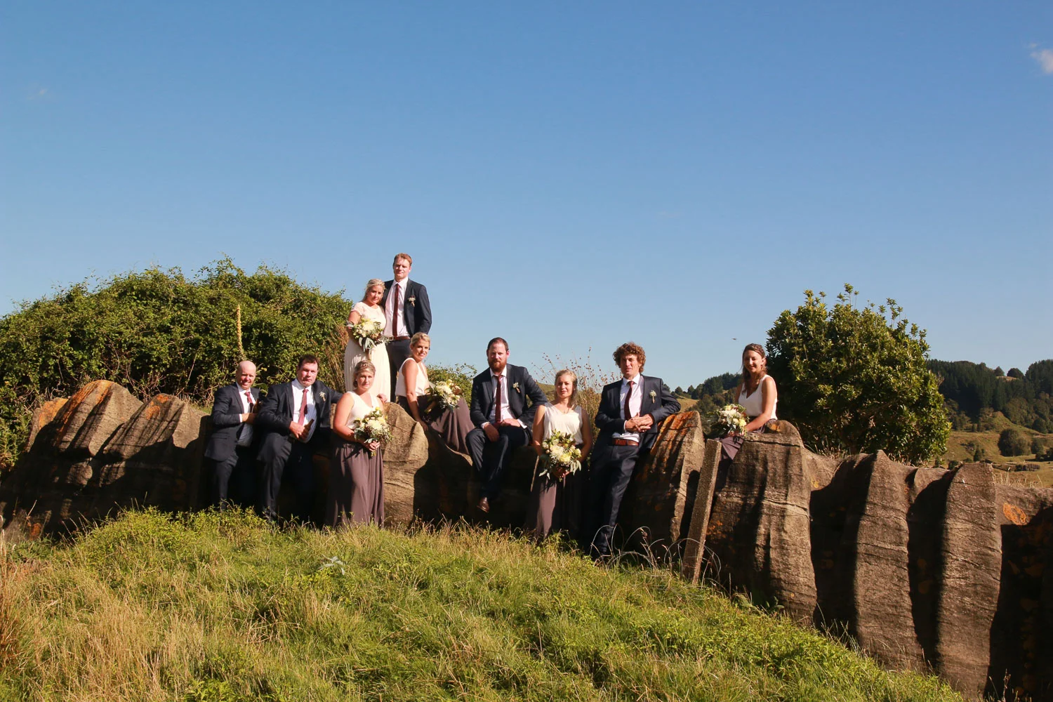 bride_and_groom_on_wedding_day_bridesmaids_and_groomsmen_all_the_wedding_party_together_leaning_on_limestone_rocks_on_farm_beautiful_day.jpg