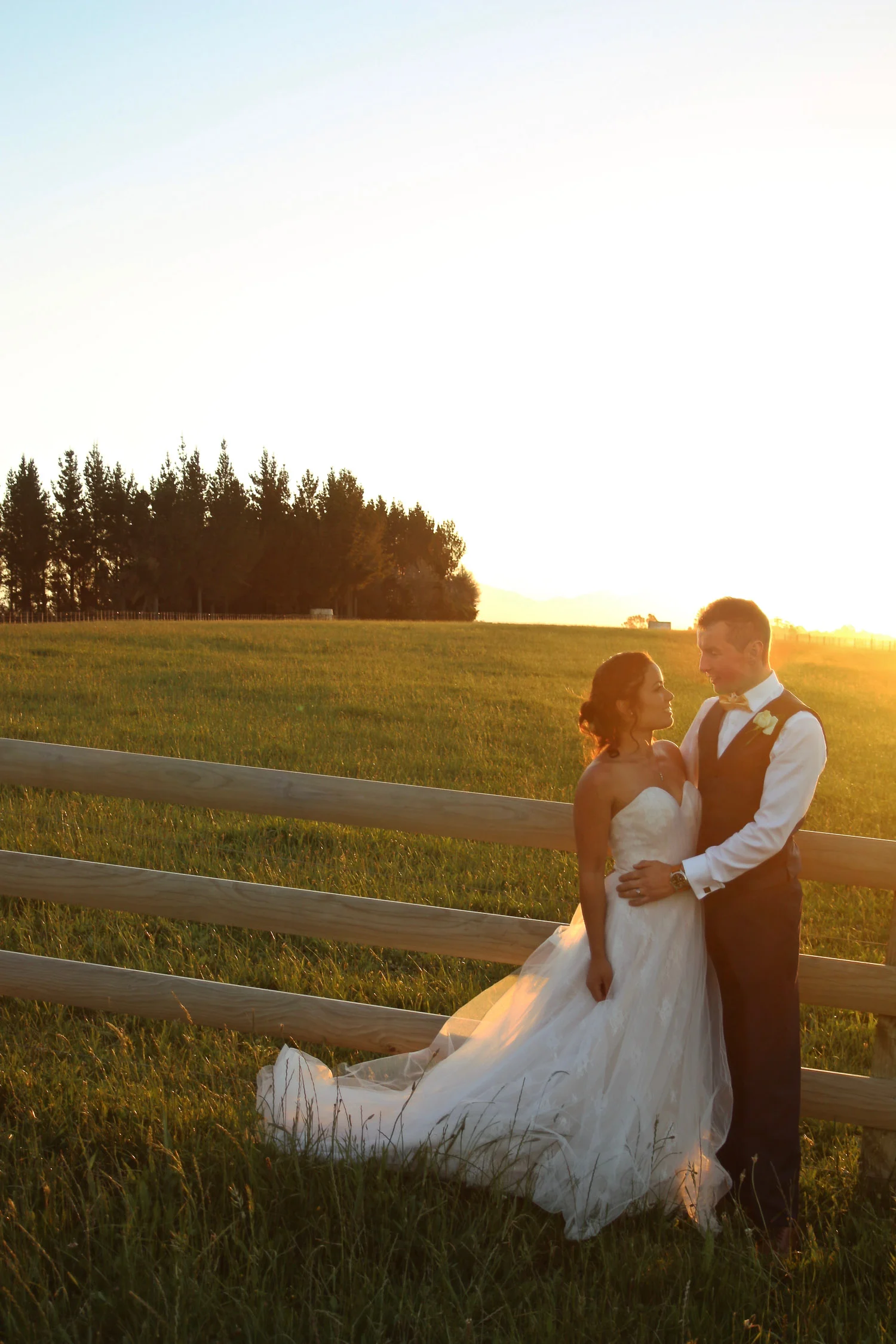 bride_and_groom_on_wedding_day_at_sunset_looking_at_eachother_in_love_by_fence_lovely_yellow_colours_trees_in_background_on_farm.jpg