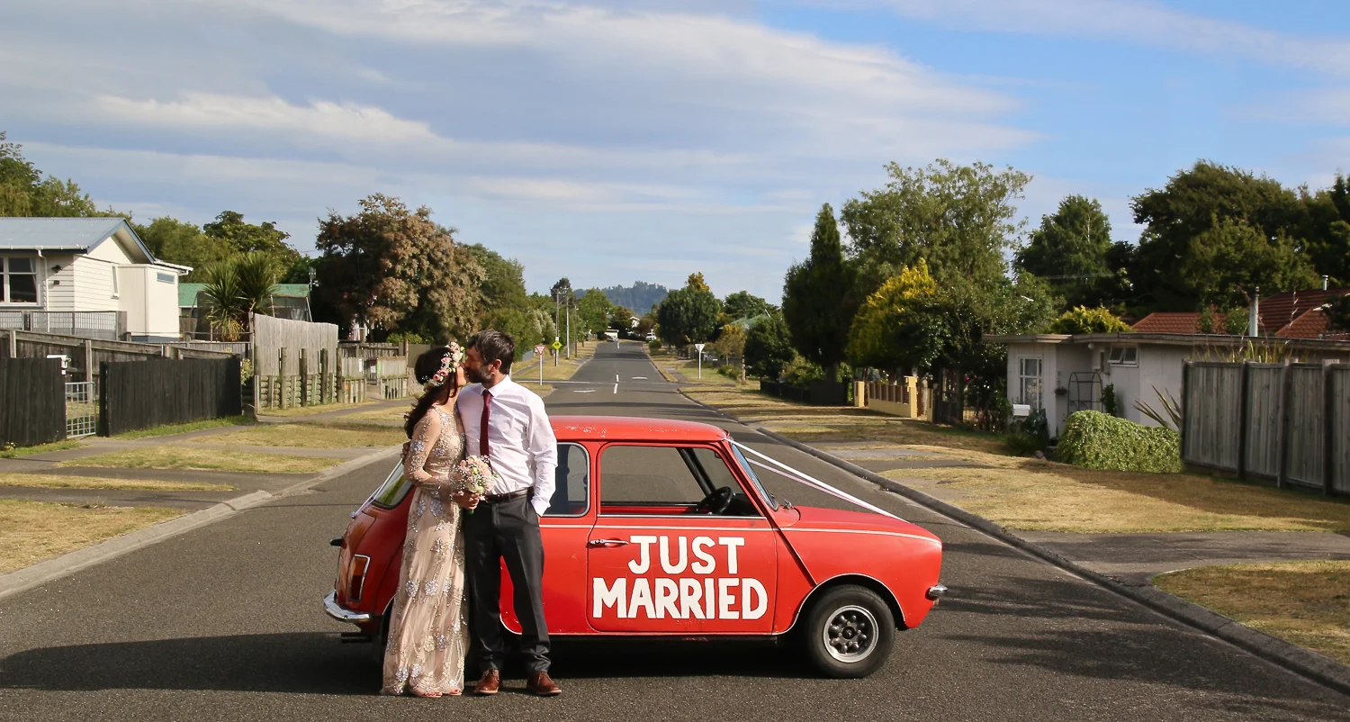 bride_and_groom_on_wedding_day_kissing_standing_in_street_next_to_red_mini_wedding_car_just_married_written_on_side_of_car.jpg