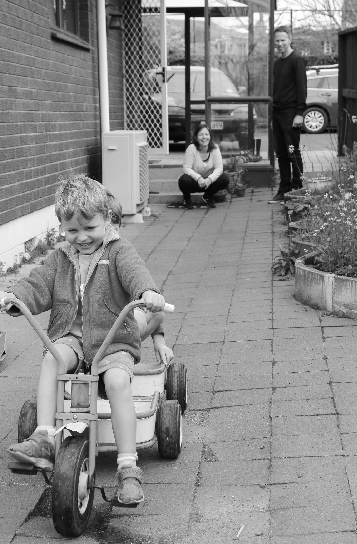 boy-playing-on-trike-mum-sitting-in-background-watching-and-smiling-black-and-white.jpg