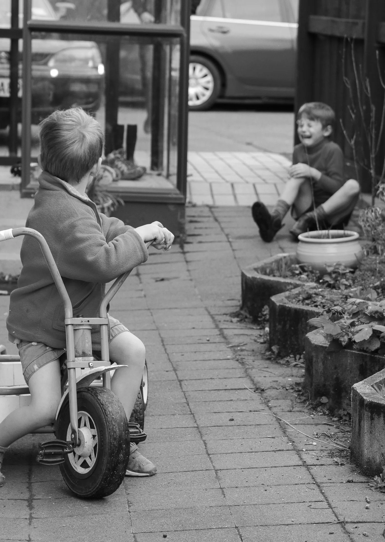 brothers-playing-together-on-trike-outside-family-home-black-and-white-one-boy-fallen-off-trike.jpg