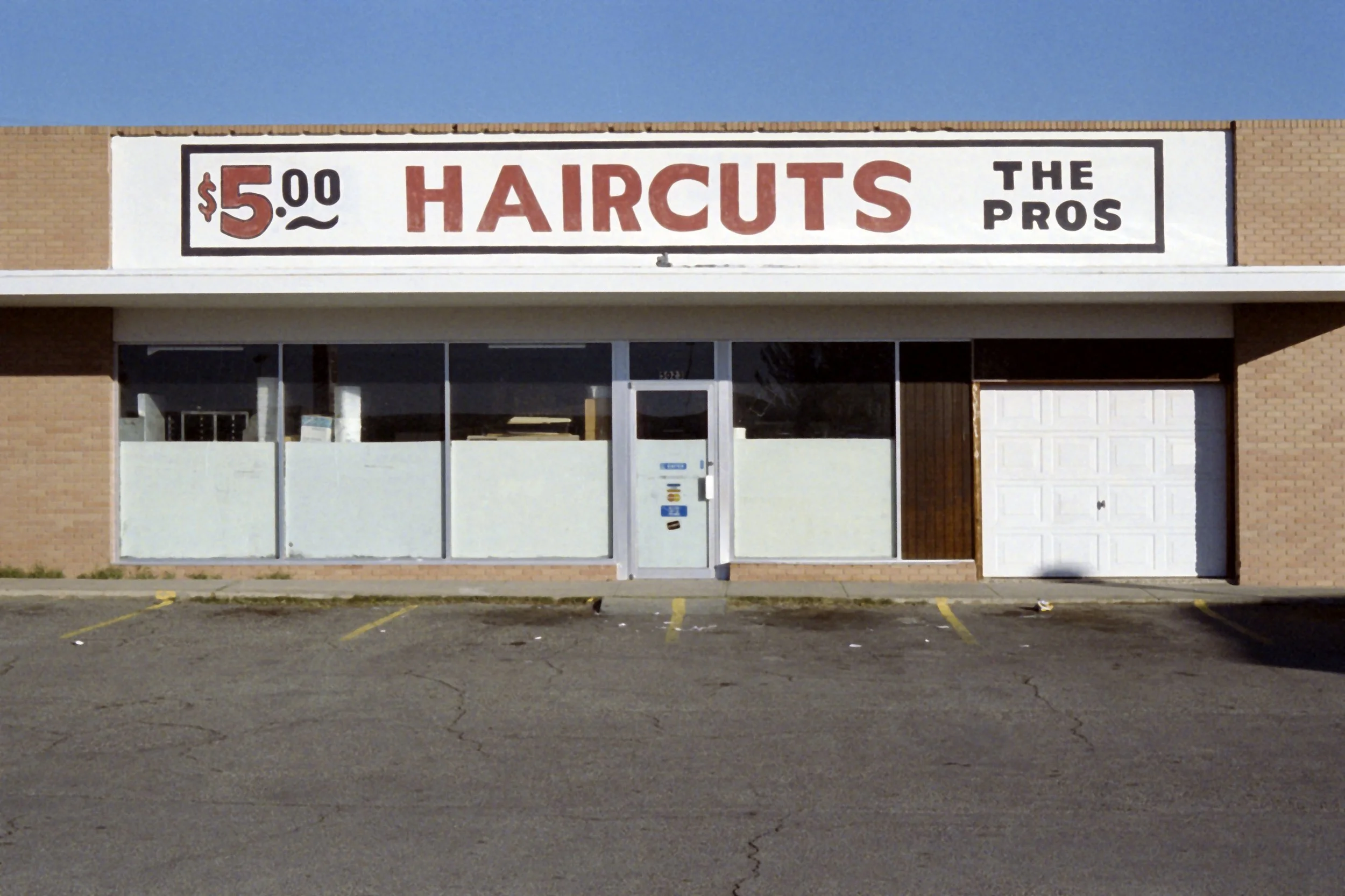 Brick building and white storefront signage with black border. Inside the border is “$5.00 Haircuts” in red text and “The Pros” in black text.