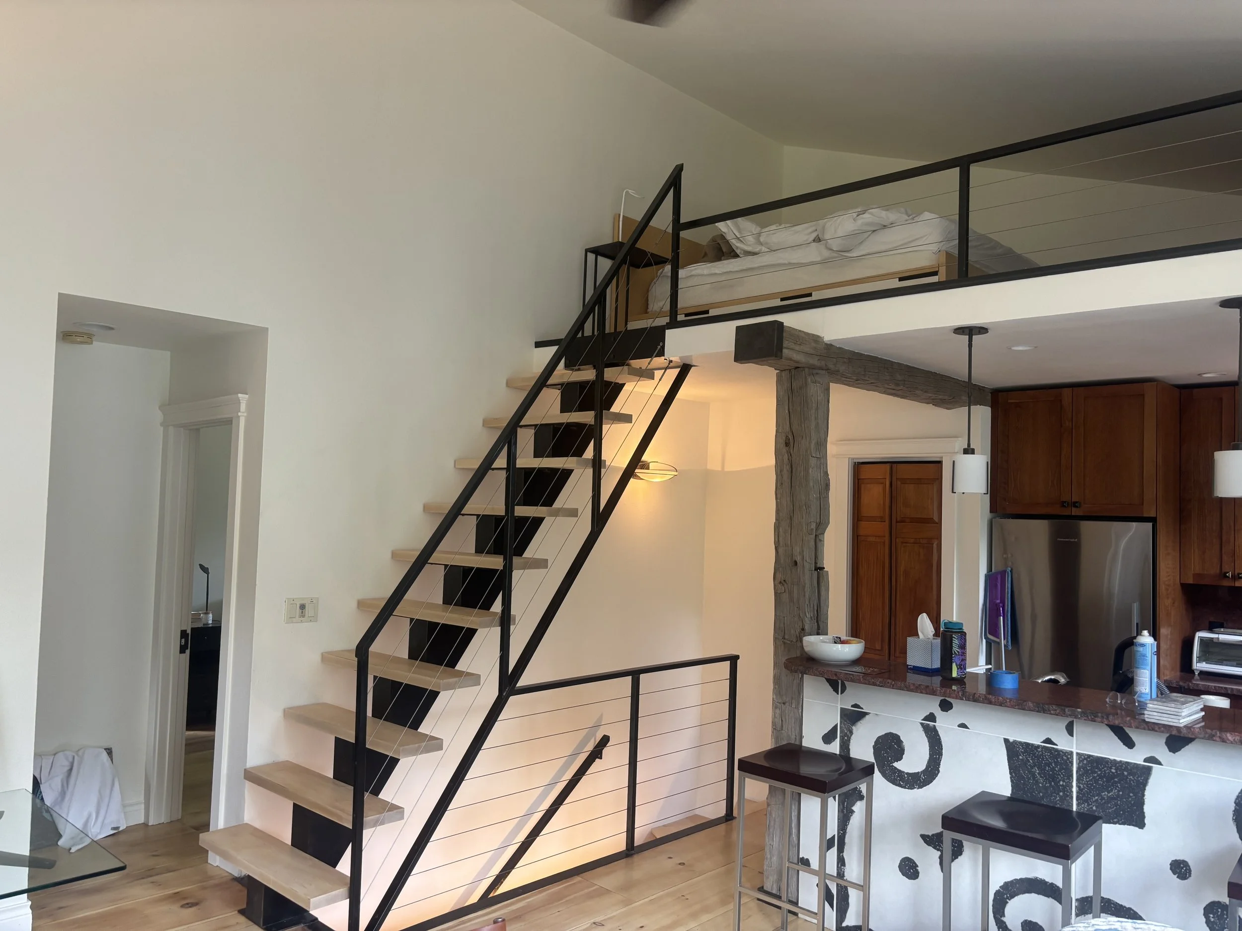 Interior view of a modern living space with a staircase leading to an upper loft area with a bed. The loft has a black metal railing, and the room features a wooden beam and kitchen cabinets in the background. There's a small round table with stools,