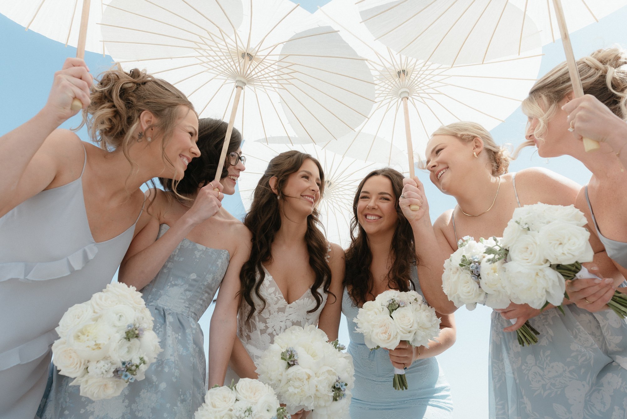 Bridesmaids laughing together holding white parasols and bouquets at Peggy's Cove Nova Scotia