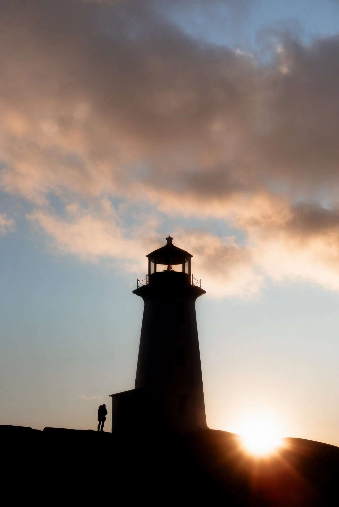 Peggy's Cove lighthouse and one lone figure silhouetted against dramatic golden sunset sky with stormy clouds 