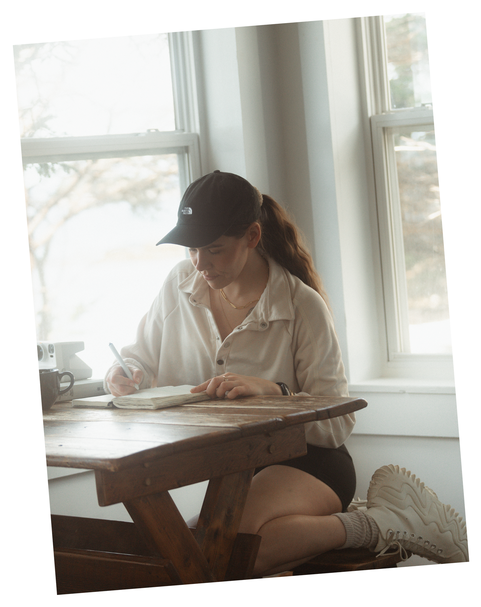 A woman sitting on a window seat, writing in a notebook on a wooden table, wearing a black cap, beige jacket, and white sneakers.