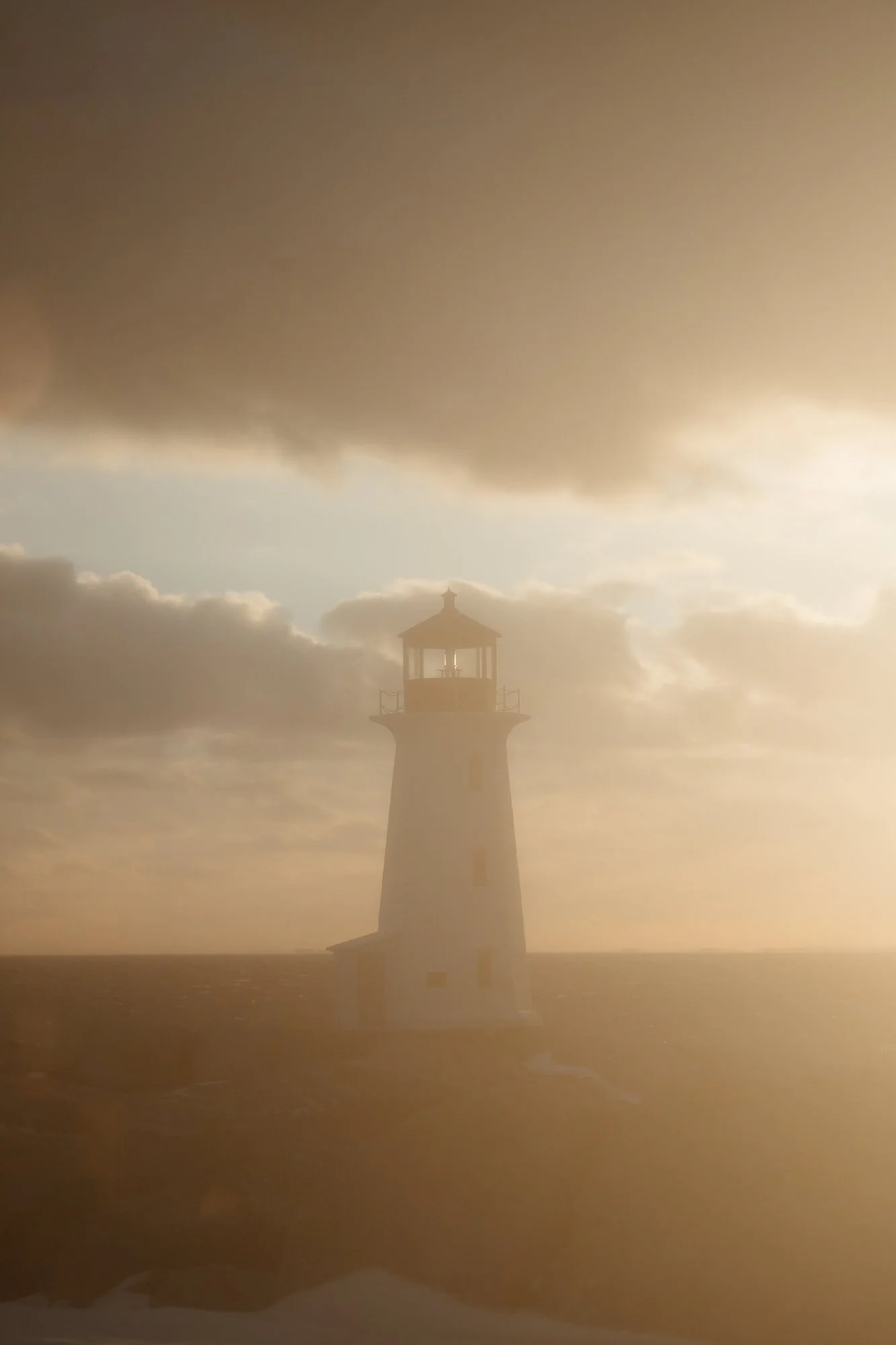 A lighthouse stands on the edge of the ocean with cloudy skies and sunlight peeking through.