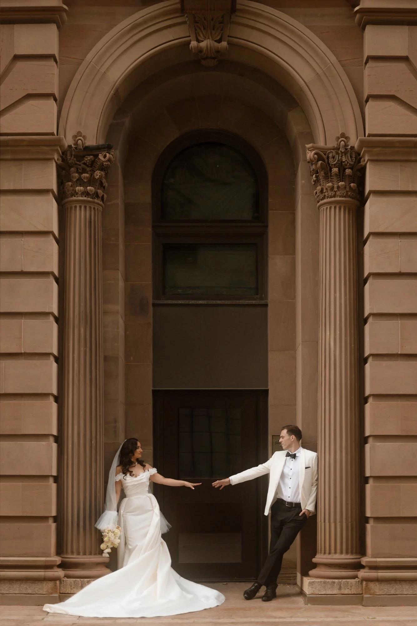 A bride and groom in wedding attire reaching out to each other in front of grand architectural columns and doorway.