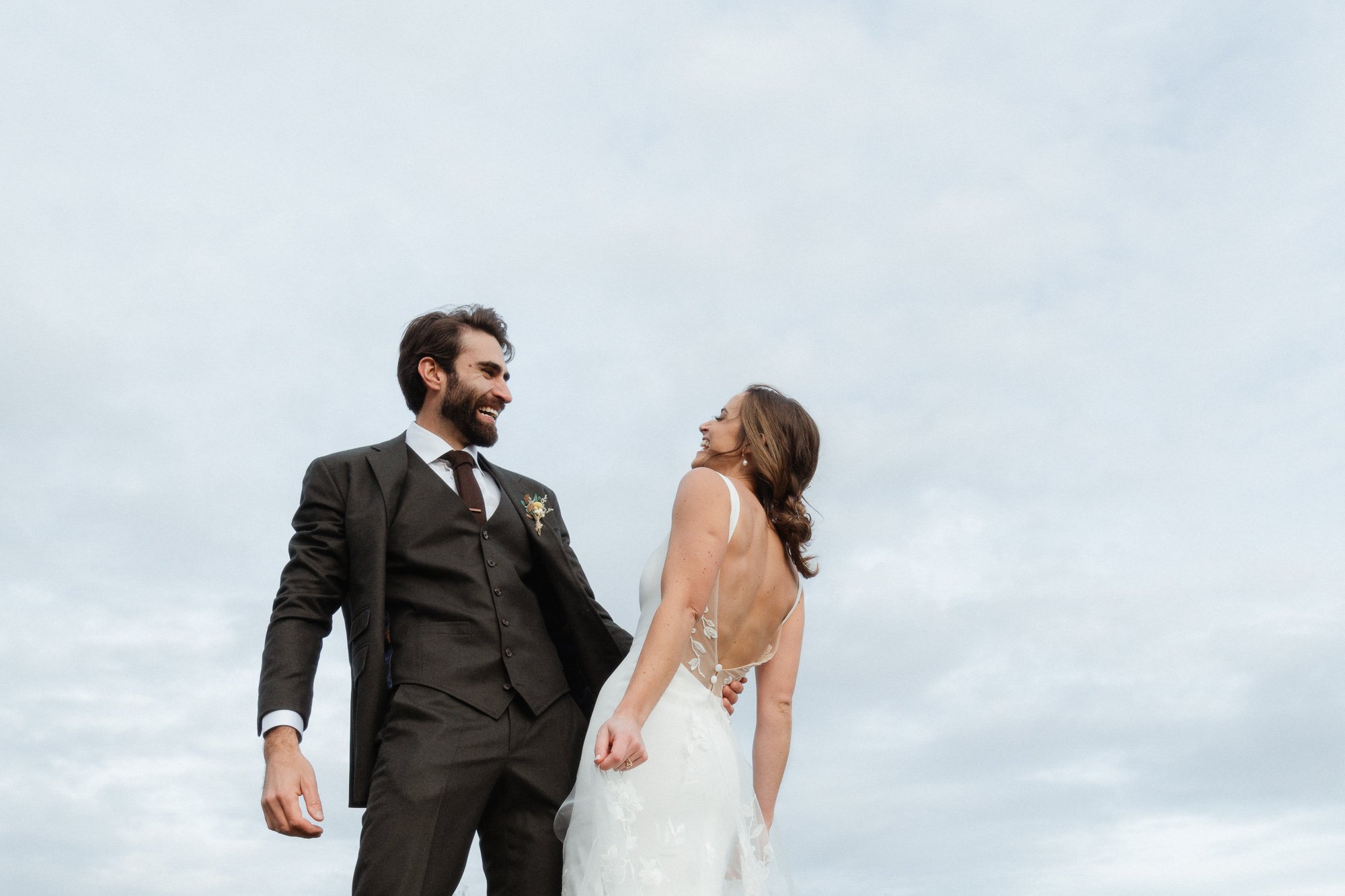 A bride and groom holding hands and smiling at each other outdoors against a cloudy sky.