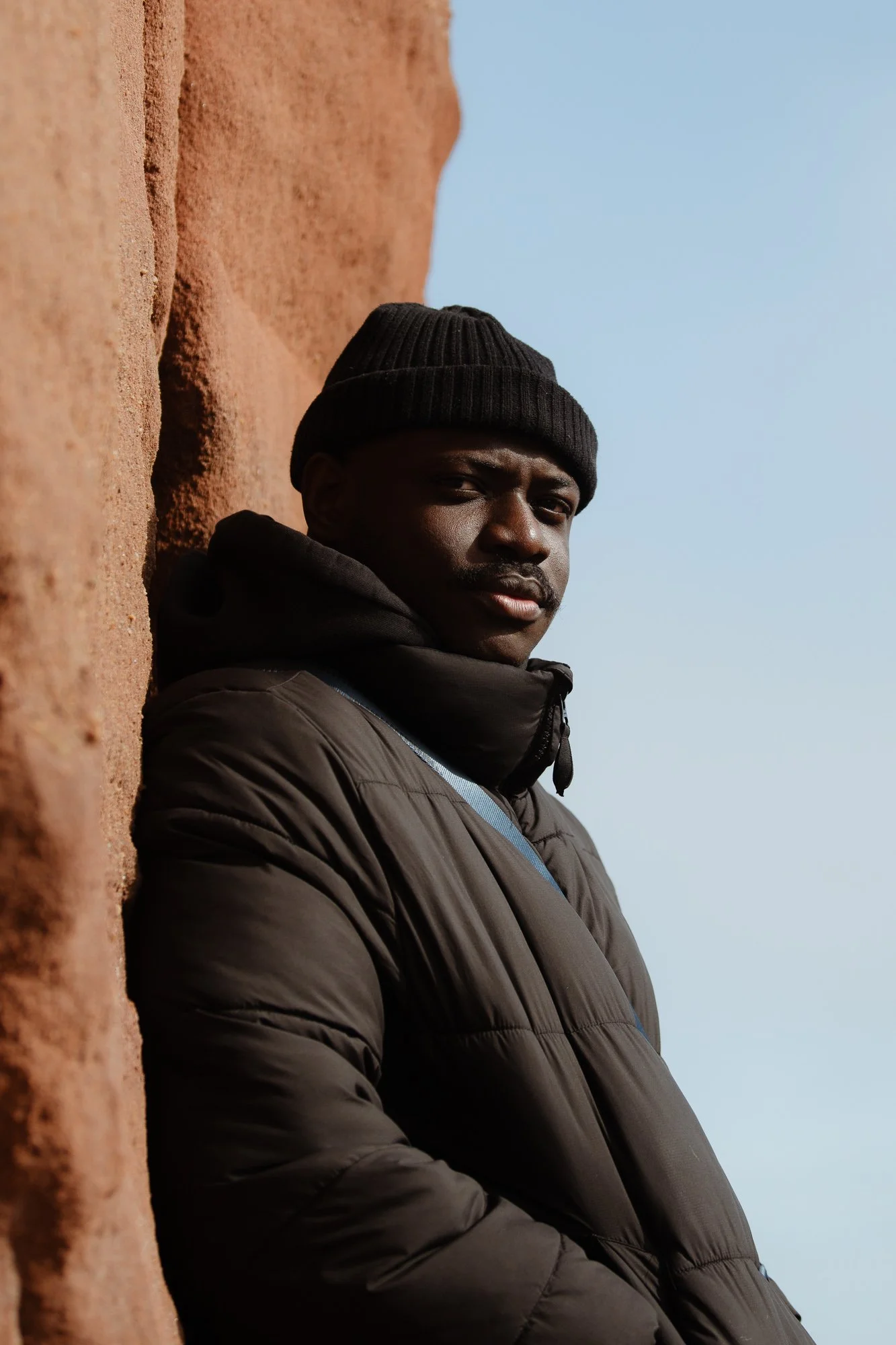 Young man wearing black beanie and puffer jacket leaning against a reddish-orange rock wall, with a clear blue sky in the background.