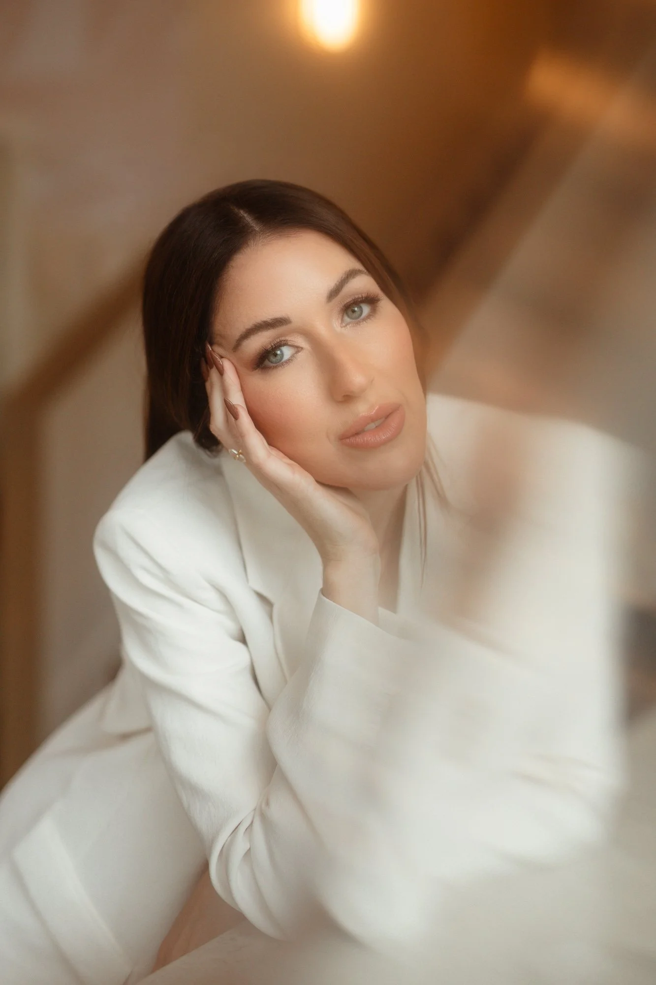 A woman with brown hair and blue eyes resting her head on her hand, wearing a white blazer, looking at the camera with a neutral expression.