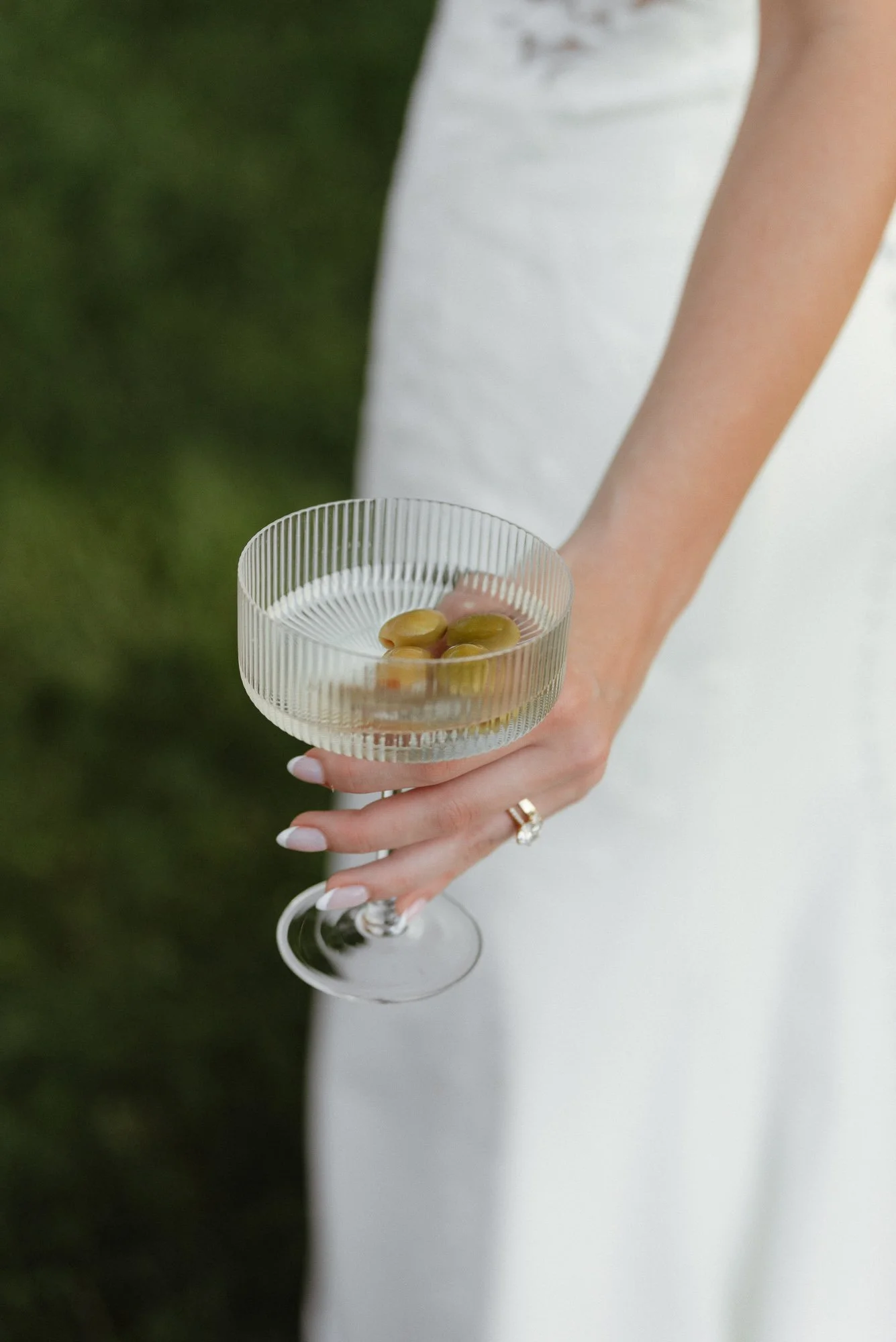 A woman's hand holding a glass dish with three green olives in water, with a blurred green background.