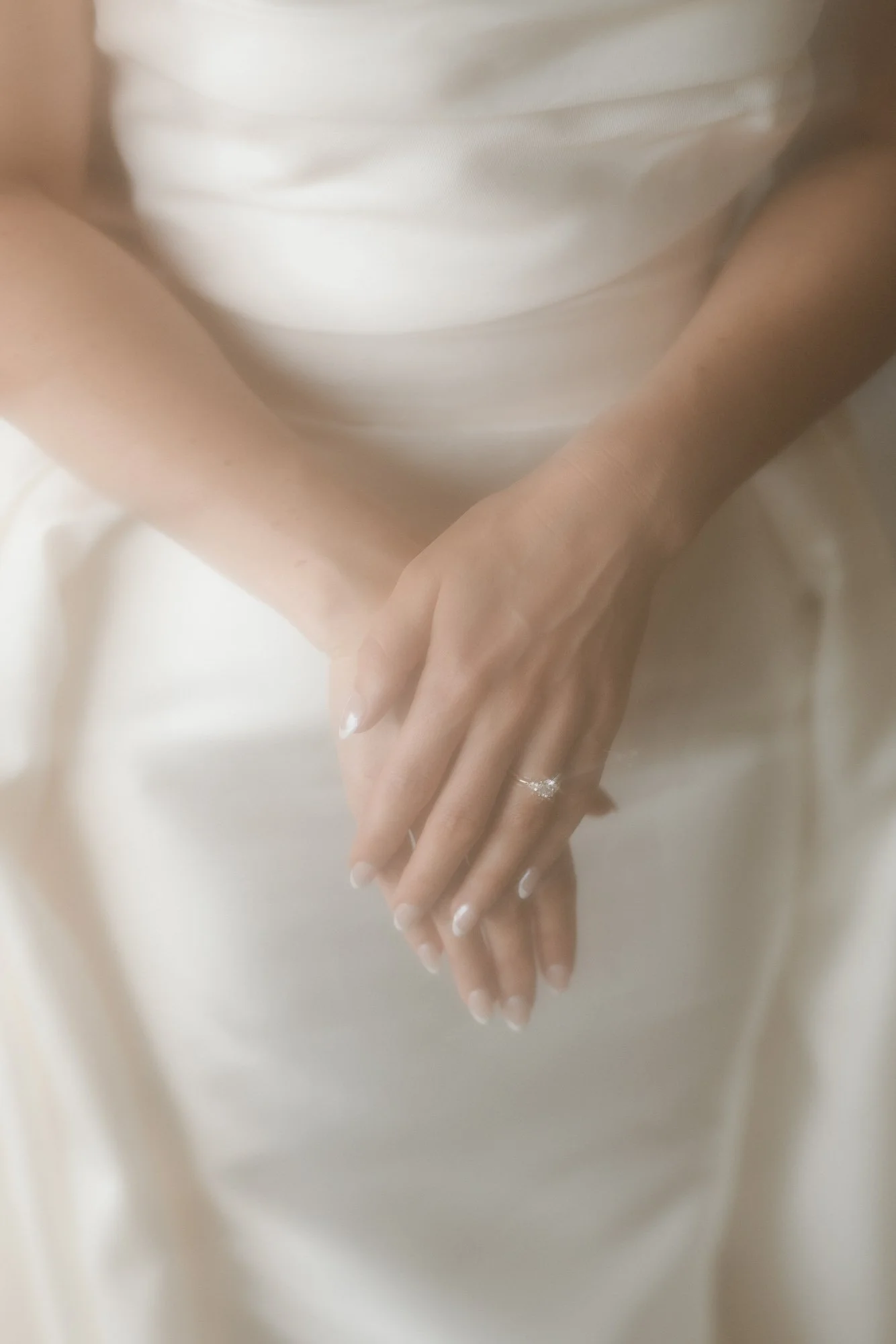 Close-up of a woman’s hands with wedding rings, resting on her lap, wearing a white dress.