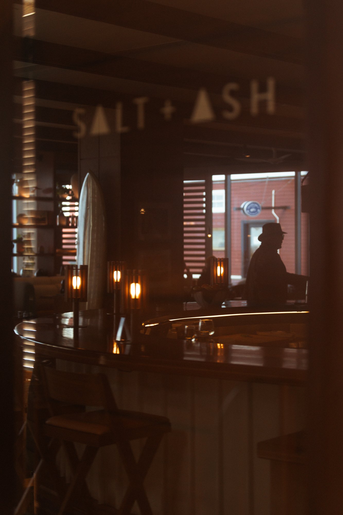 A dimly lit interior of a restaurant or bar with a silhouette of a person wearing a hat and sitting at a counter, reflected in a glass. The reflection shows a sign with the text "Salt + Ash" and some warm lighting fixtures.
