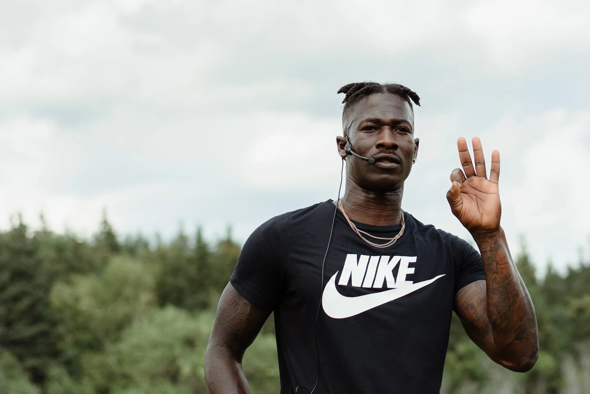 A young man running outdoors, wearing a black Nike t-shirt and a headset, making an OK sign with his right hand, with a background of trees and cloudy sky.