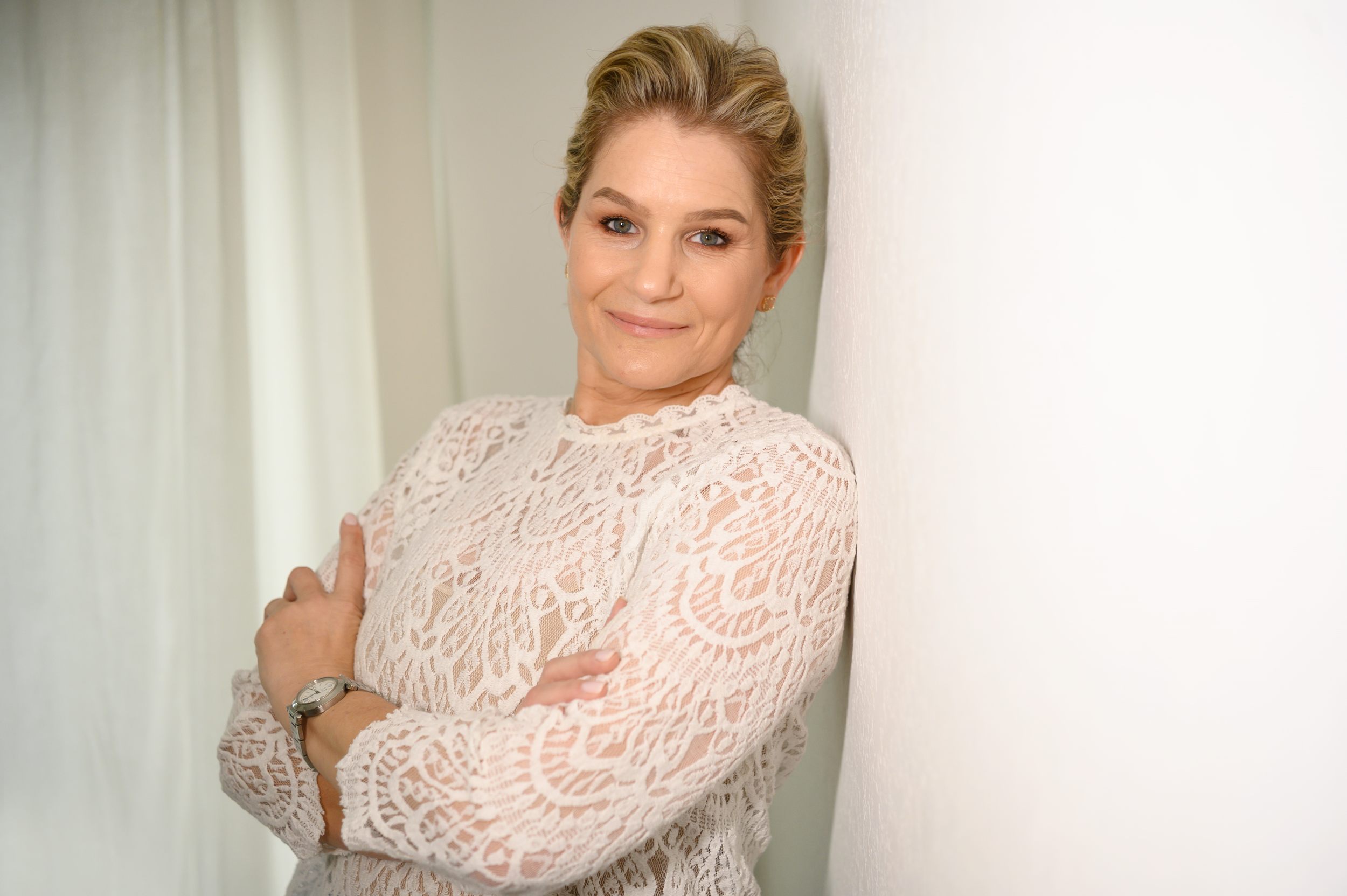 A women with blonde hair, leaning against a wall with arms crossed, smiling at the camera.