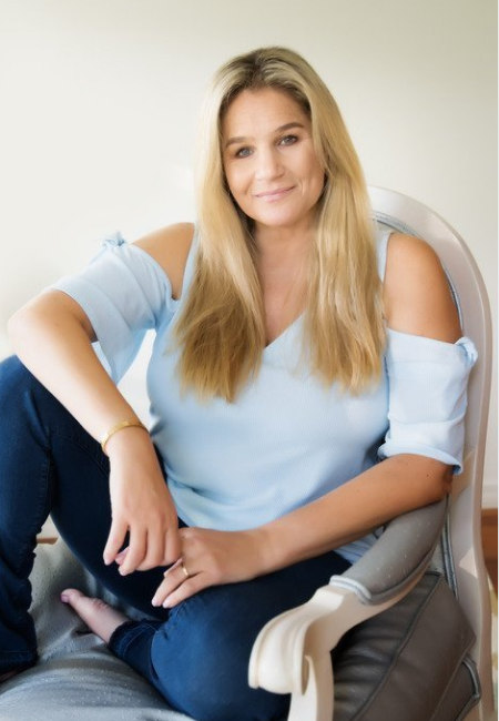 A woman with long blonde hair wearing a light blue top sits on a gray armchair, smiling at the camera.