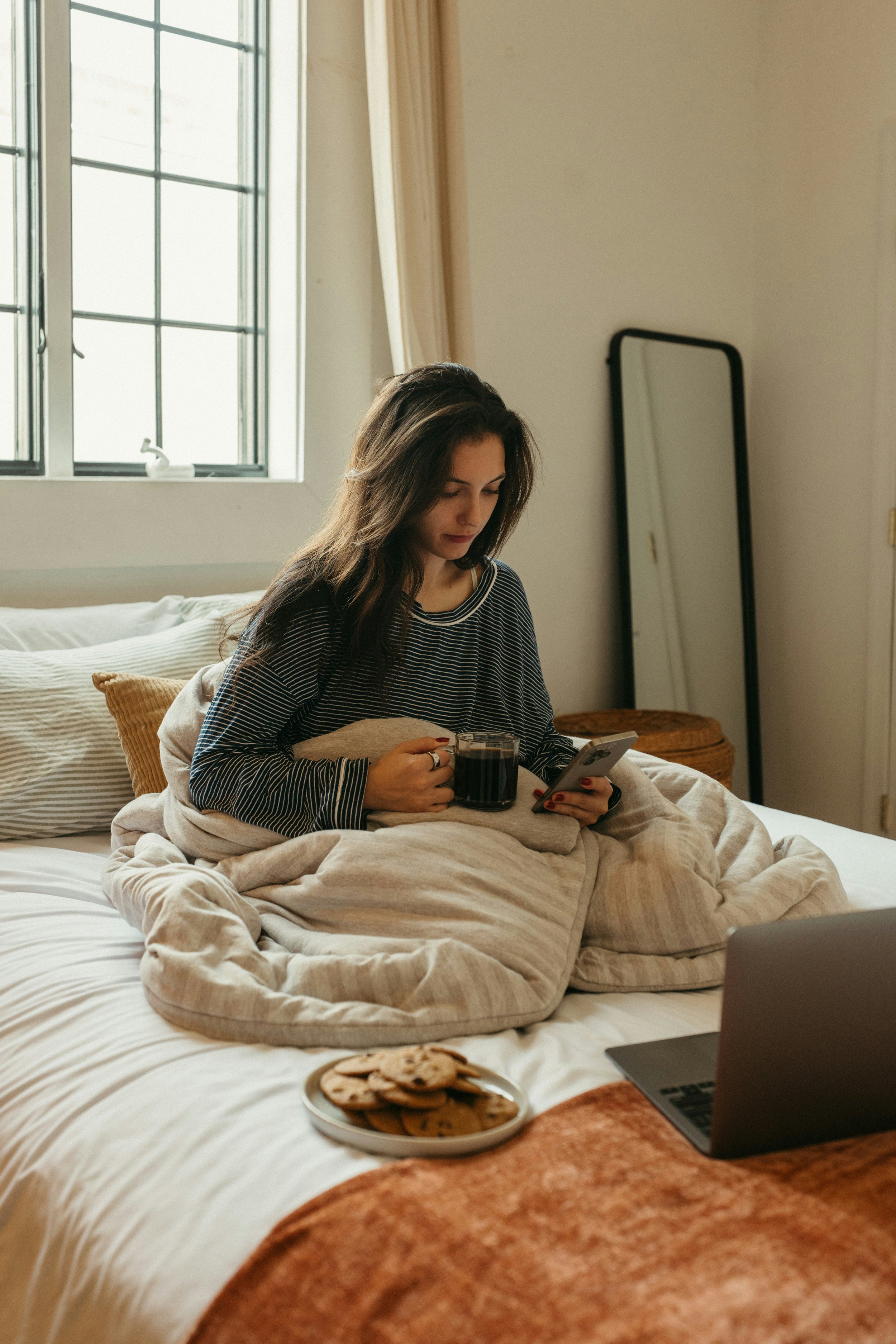 Woman sitting on bed, looking at phone while holding a coffee | Conflicted