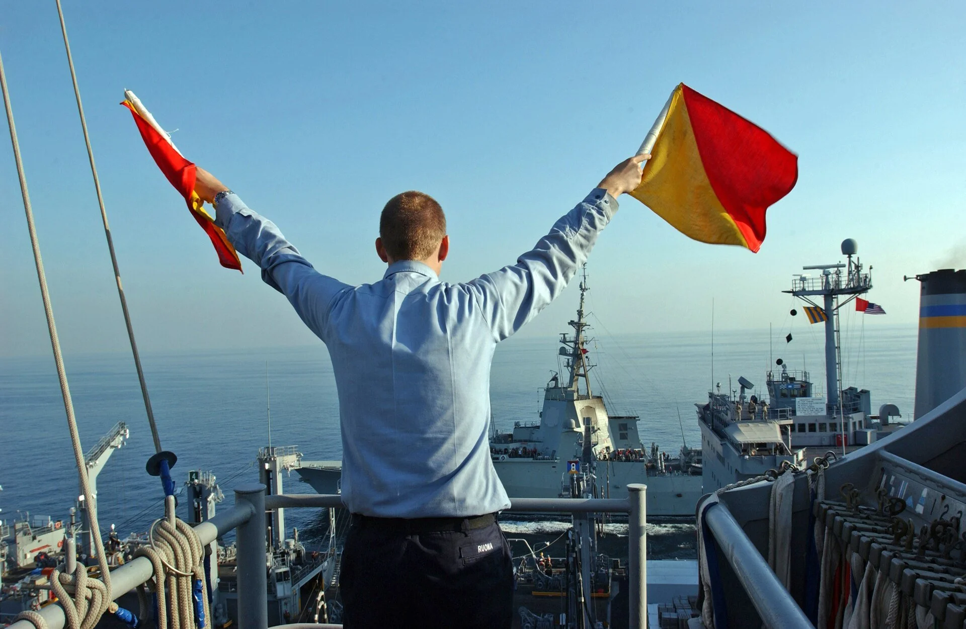 A sailor on a ship holding two red-and-yellow flags up at 45-degree angles