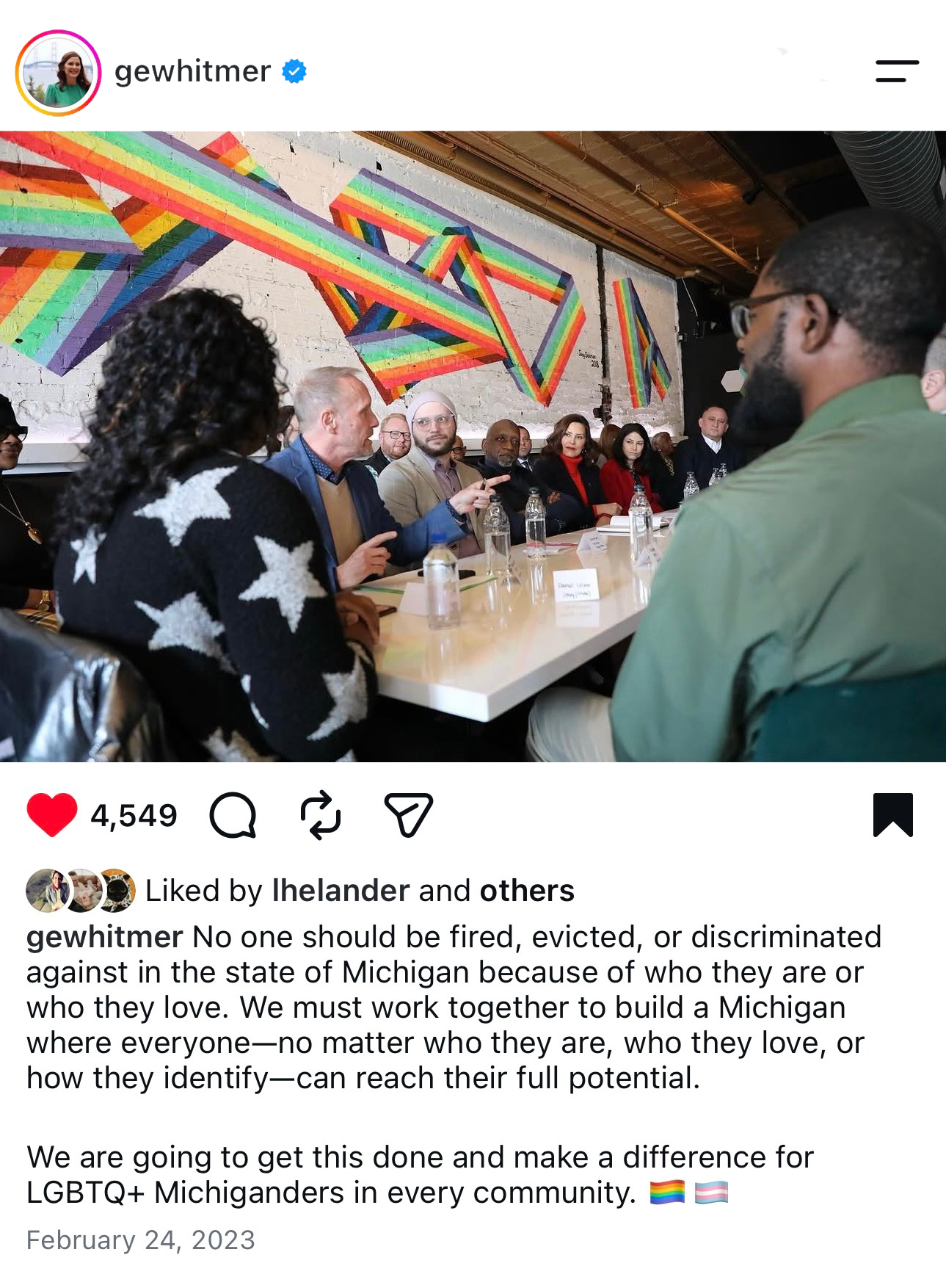 A diverse group of people attending a panel discussion or meeting in a modern, artsy conference room with rainbow-colored artwork on a white brick wall.
