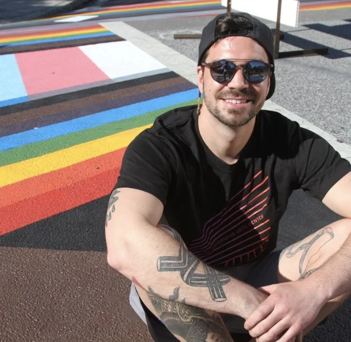 Man with tattoos wearing sunglasses and a black cap, sitting on a colorful rainbow crosswalk in the sunshine.