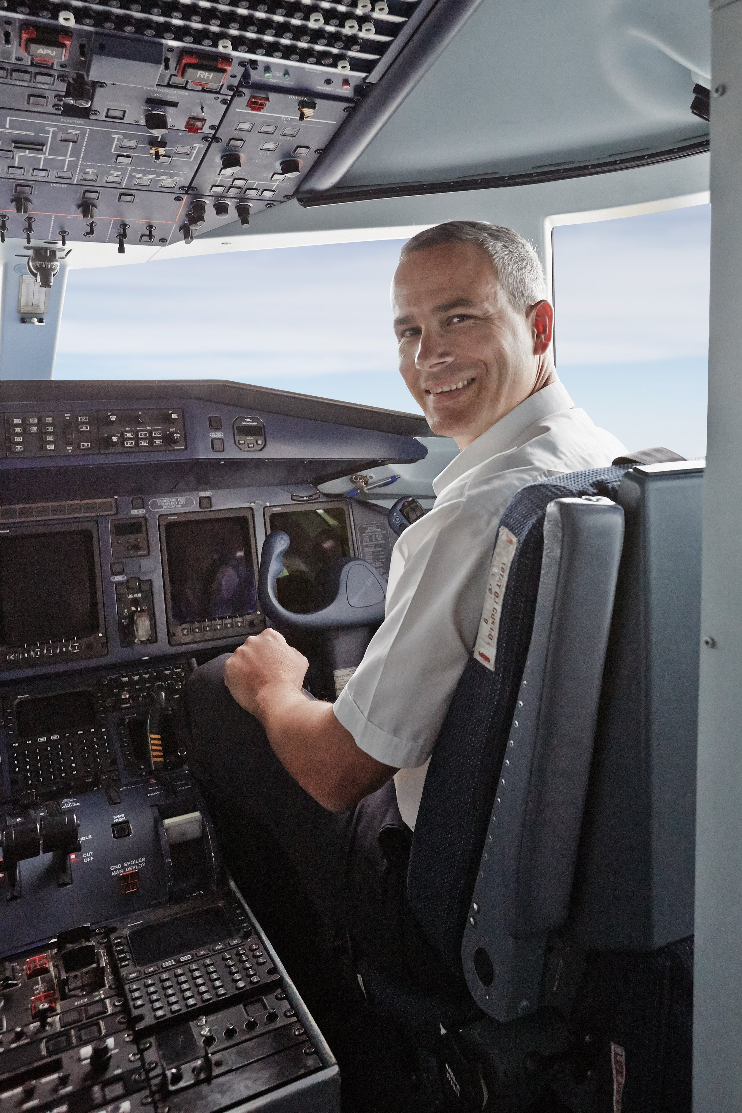 Ein Pilot sitzt im Cockpit eines Flugzeugs, schaut lachend in die Kamera und trägt eine weiße Uniform. Das Cockpit ist mit Vielzahl von Instrumenten, Anzeigen und Steuerungen ausgestattet.