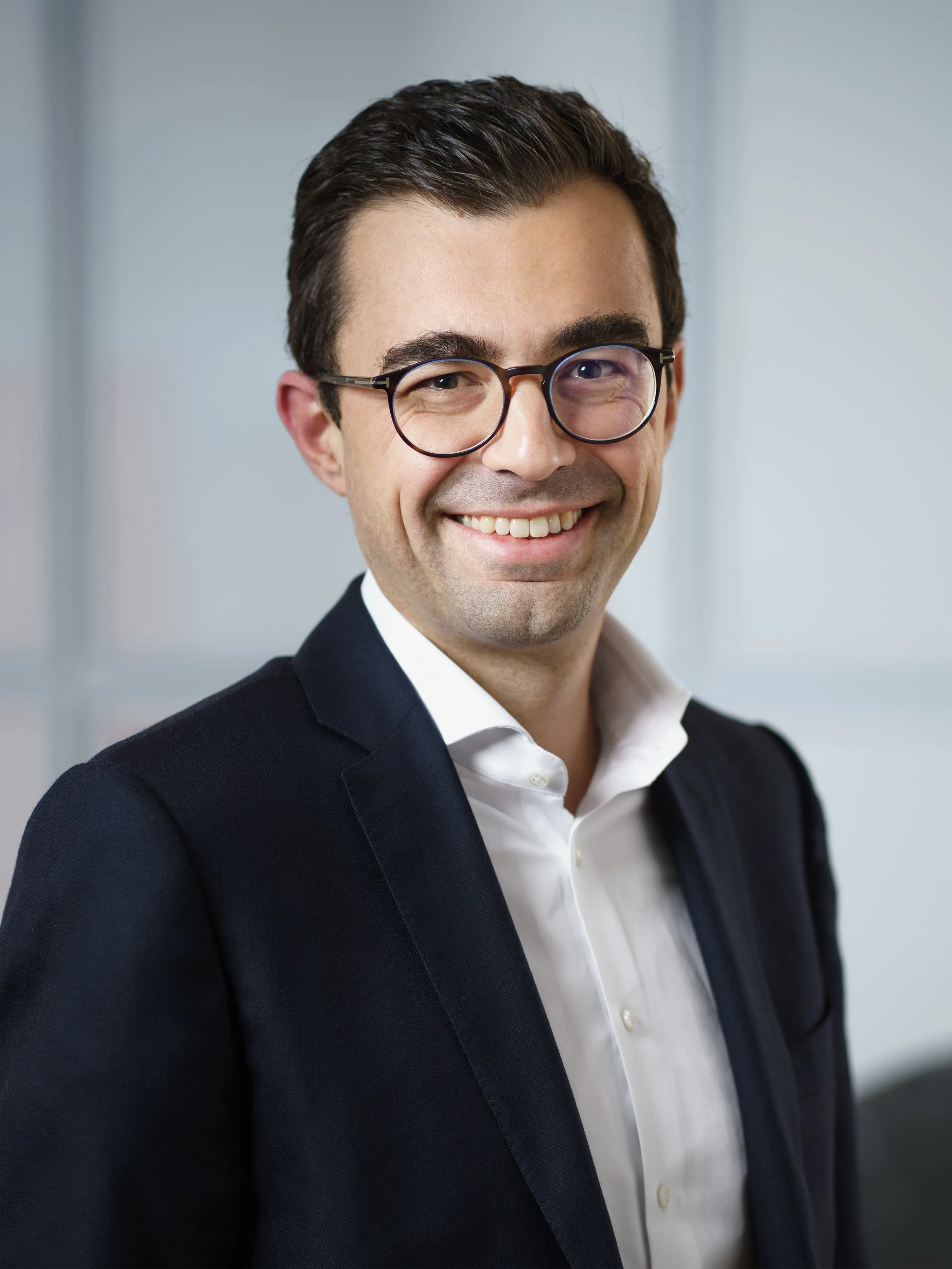 Ein lächelnder Mann in einem dunklen Anzug und einer weißen Hemd, der eine Brille trägt. Der Hintergrund ist unscharf grau.