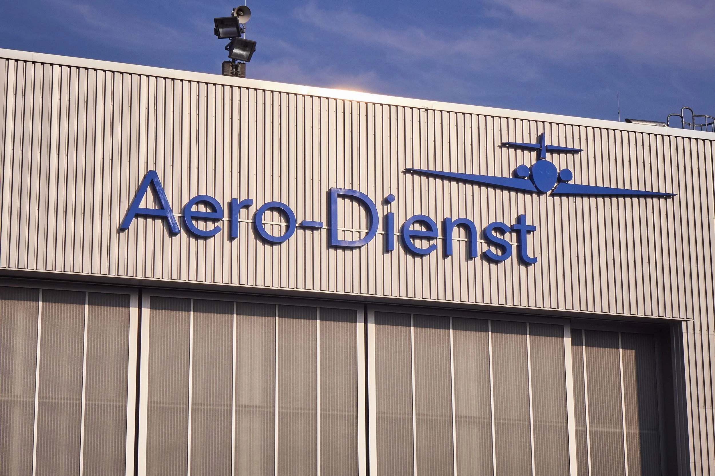 Das Bild zeigt das Gebäude der Aero-Dienst mit blauer Schrift und einem blauen Flugzeuglogo an der Fassade, unter einem blauen Himmel mit einigen Wolken.