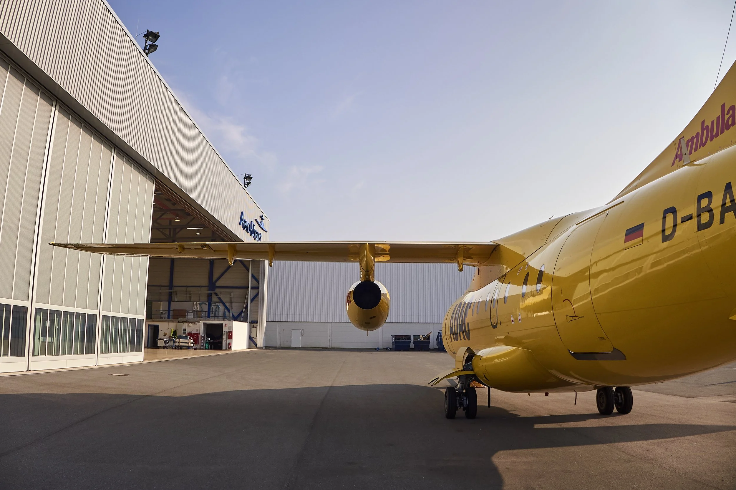 Ein gelbfarbenes Flugzeug vor einem Hangar mit der Aufschrift AeroDients, im Hintergrund ist das Logo eines Flugsicherheitsdienstleisters sichtbar.