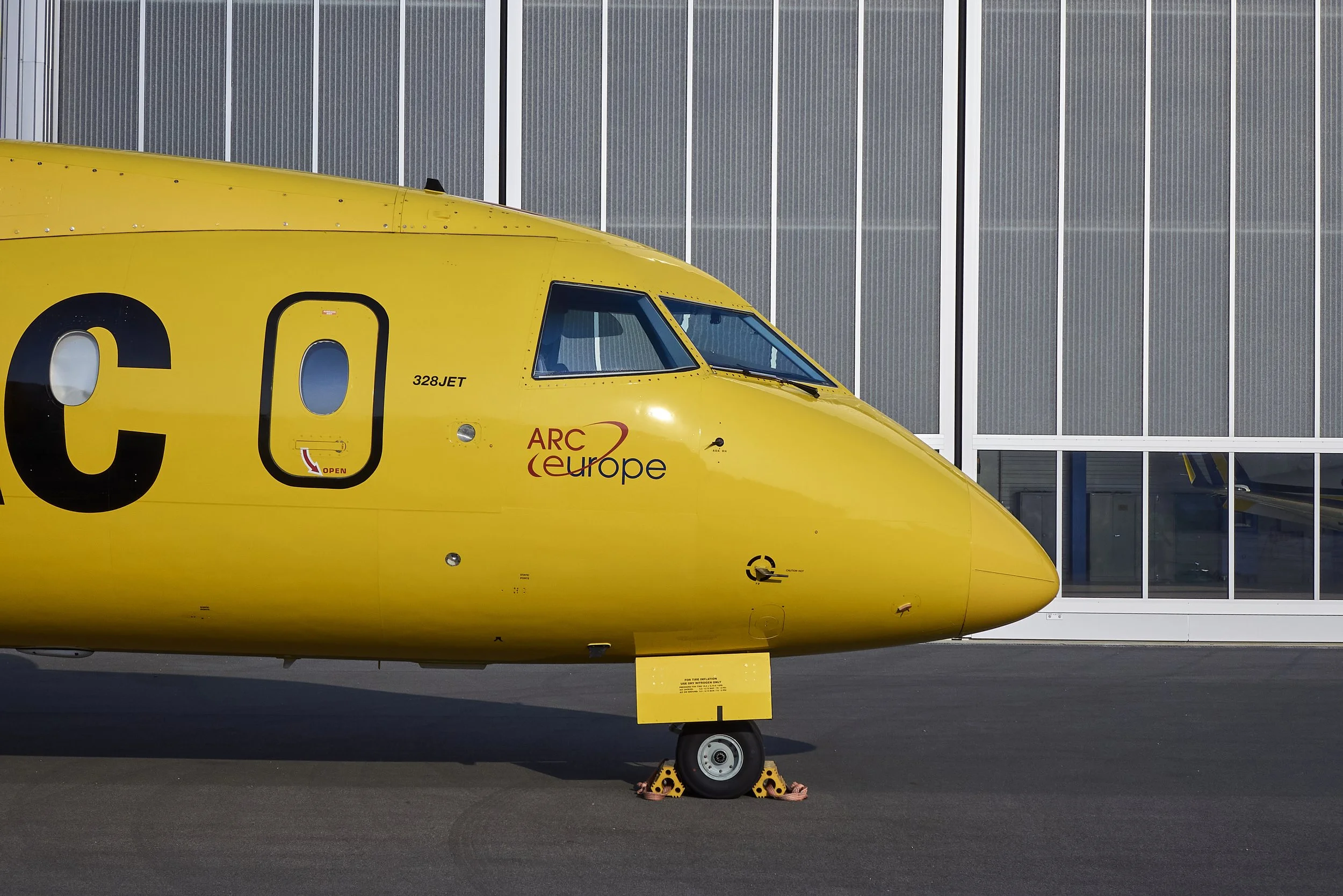 Ein gelber Flugzeugrumpf mit dem Schriftzug ARC Europe und der Nummer 328JET, vor einer modernen Gebäudefassade mit großen Fenstern.
