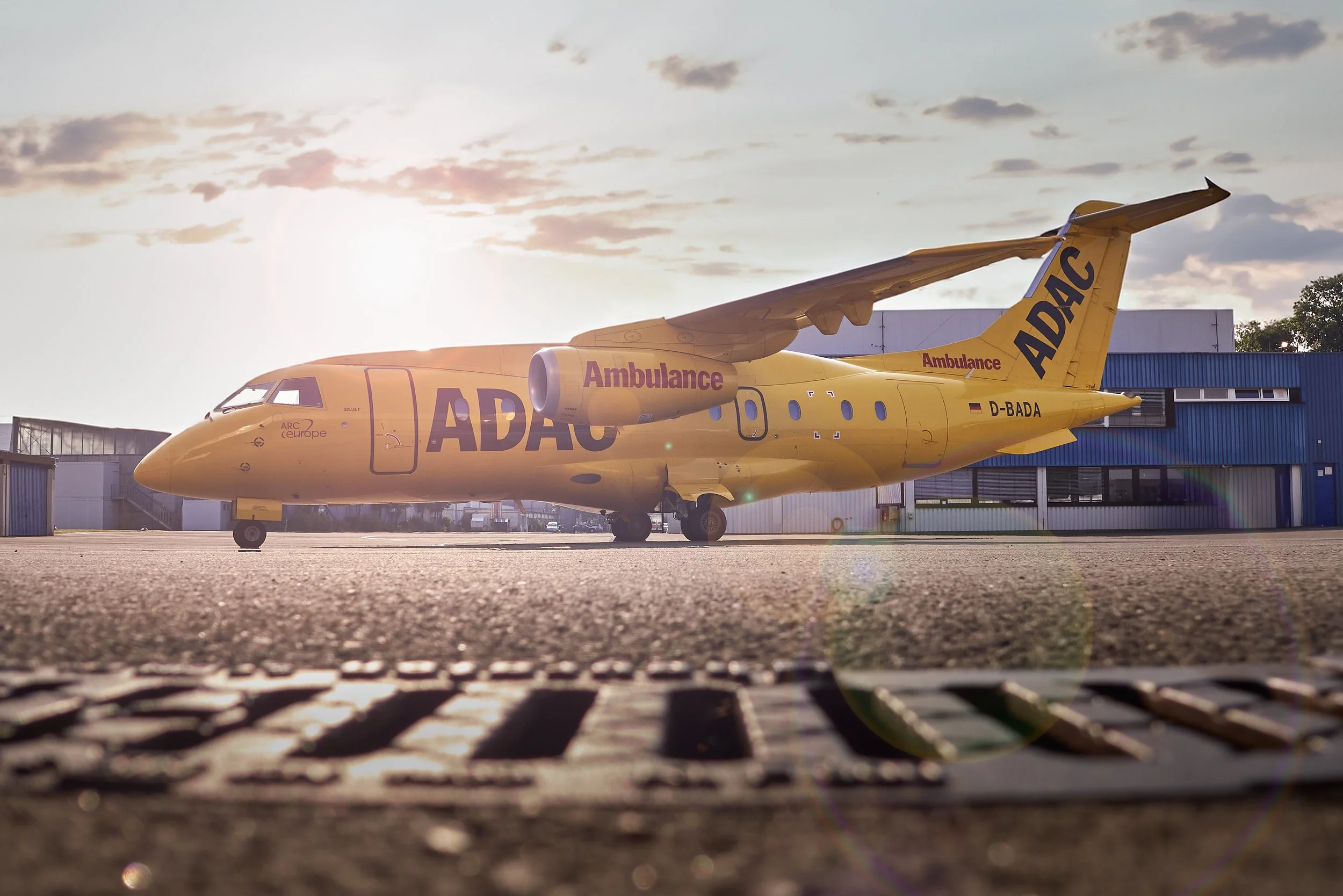 Ein gelbes Flugzeug, das als Ambulanz gekennzeichnet ist, steht auf dem Flughafen bei Sonnenuntergang, mit einem blauen Gebäude im Hintergrund.