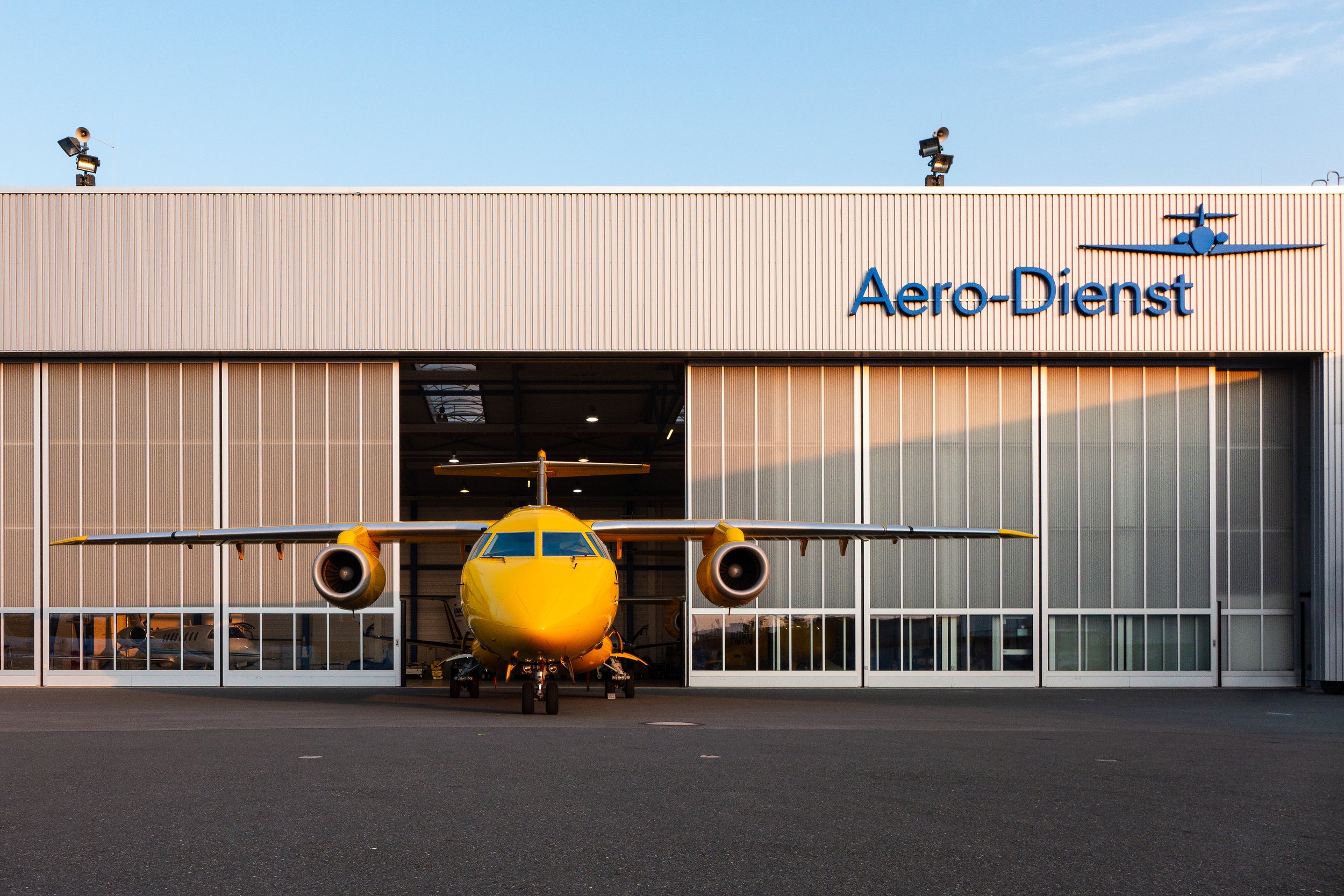 Ein gelbes Flugzeug steht vor einem Hangar mit der Aufschrift 'Aero-Dienst'.