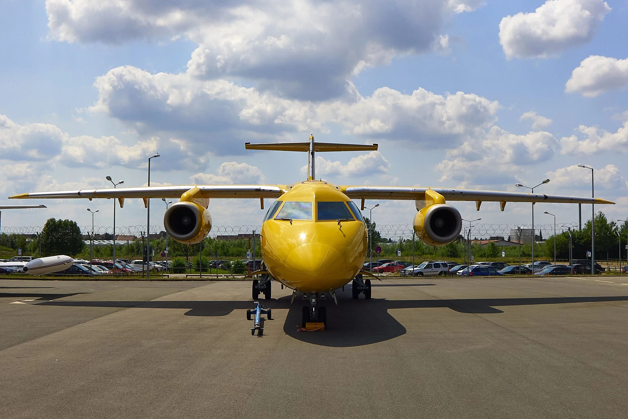 Blick auf eine gelbe Flugzeugmitte auf einem Flughafen, im Hintergrund die Parkplätze mit Autos und ein Himmel mit Wolken.