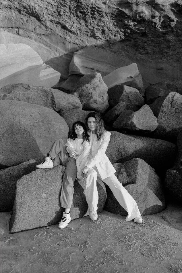 Two women, one young girl, sitting on large rocks in a barren, rocky landscape. The women are dressed in light-colored clothing, with the woman on the right in a blazer and pants, and the girl is smiling, wearing T-shirts and sneakers.
