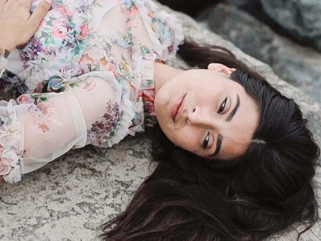 A woman with long dark hair lying on the ground, wearing a floral dress with ruffles.