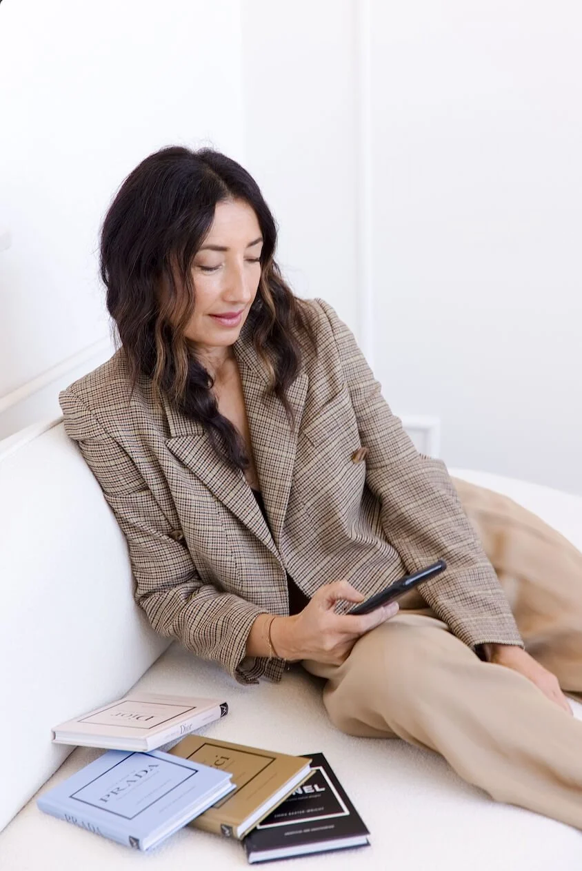 A woman with wavy brown hair wearing a beige checkered blazer sitting on a white sofa, looking at her phone. Several fashion magazines are spread out on the sofa next to her.