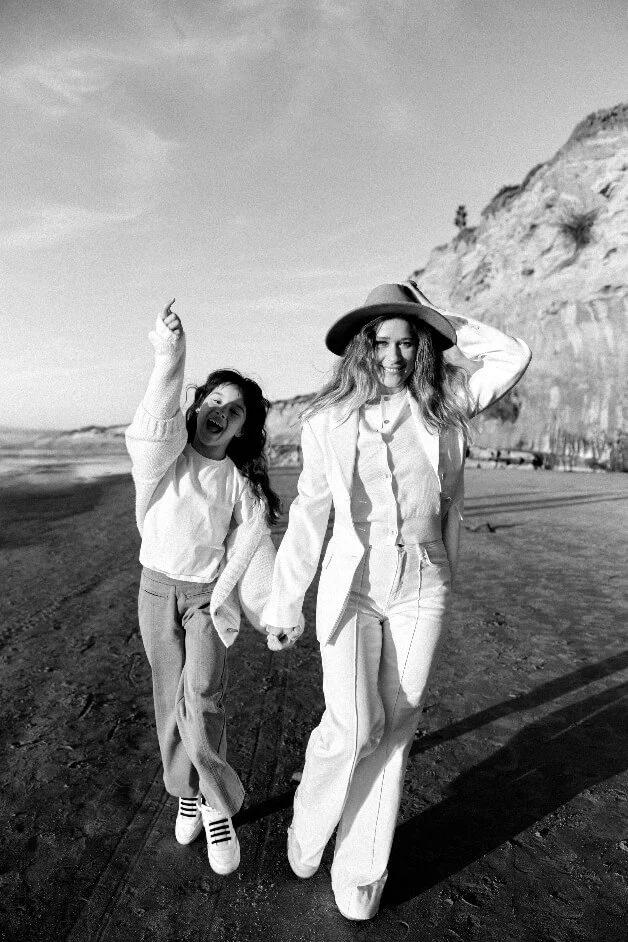 Two women walking on a beach holding hands, one smiling and wearing a hat, the other excited with finger pointing up, cliffs in background, black-and-white photo.