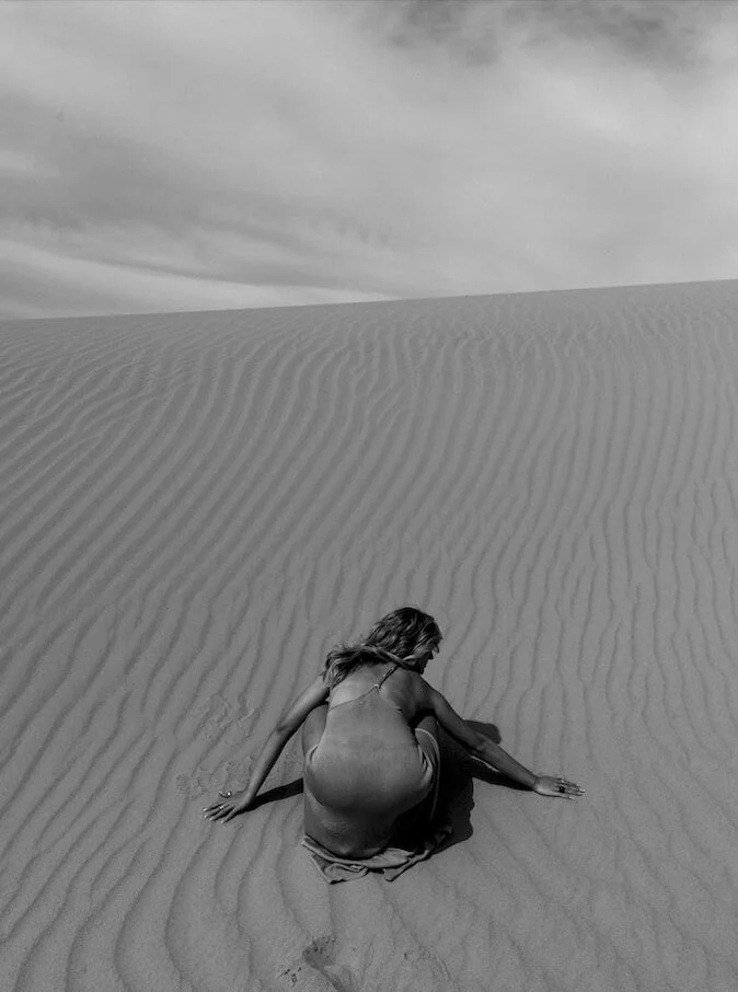 portrait in black and white of a woman bending down on the sand at the sand dunes