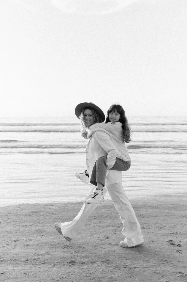 A woman carrying a young girl on her back walking on the beach, with ocean waves in the background, in black and white.