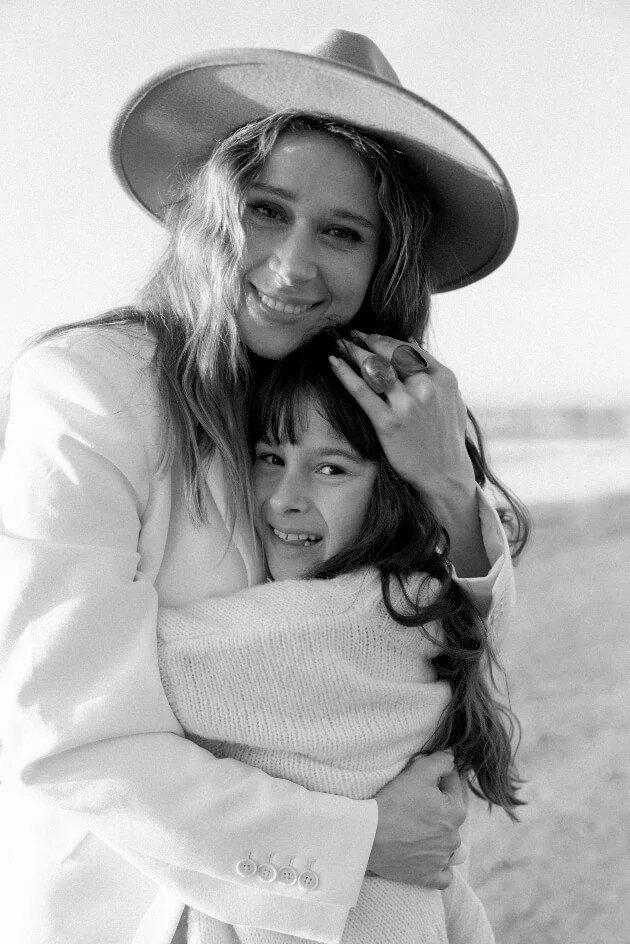A woman and a girl hugging on the beach, smiling at the camera. The woman is wearing a hat and a light-colored jacket, and the girl has long hair and is wrapped in a sweater.