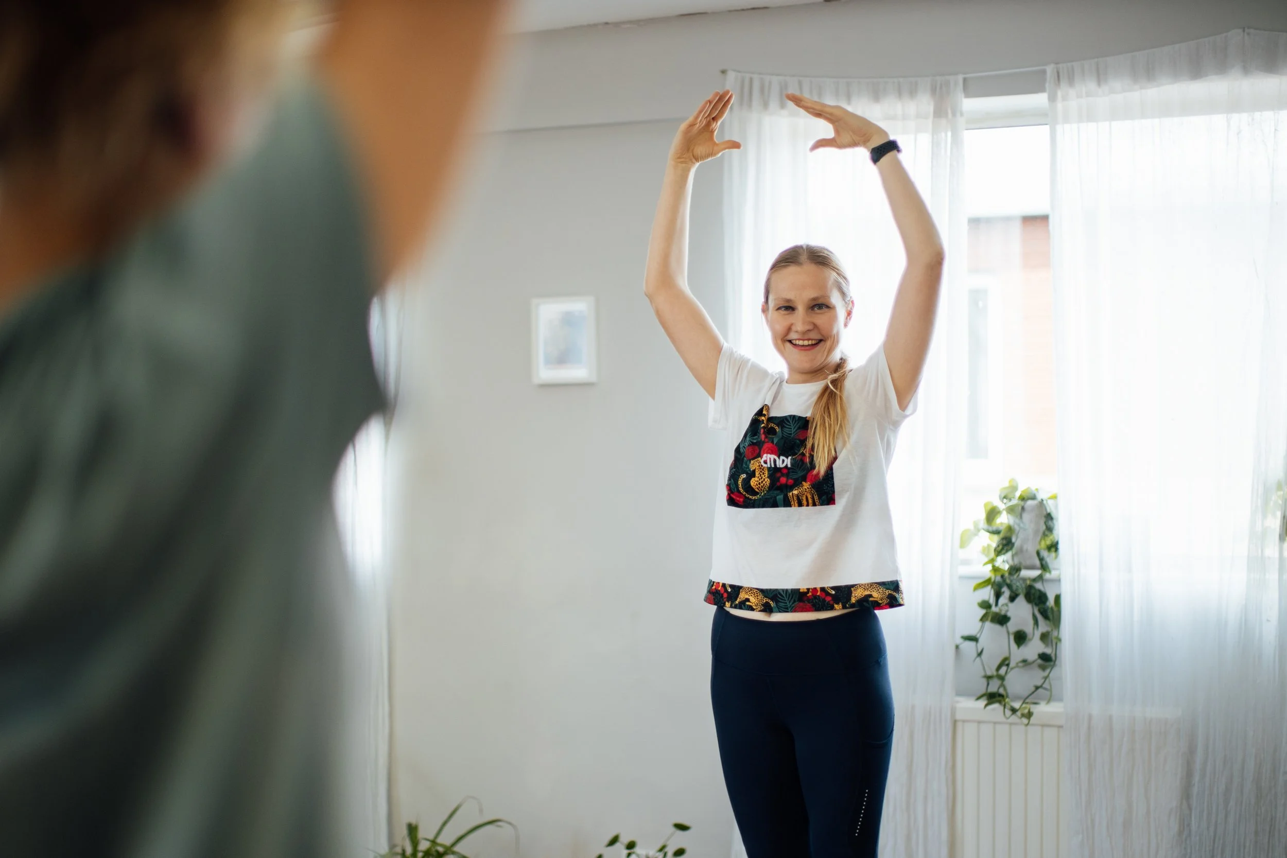  Smiling Pilates instructor standing in a bright studio with arms lifted overhead in a graceful movement, demonstrating posture and alignment. 