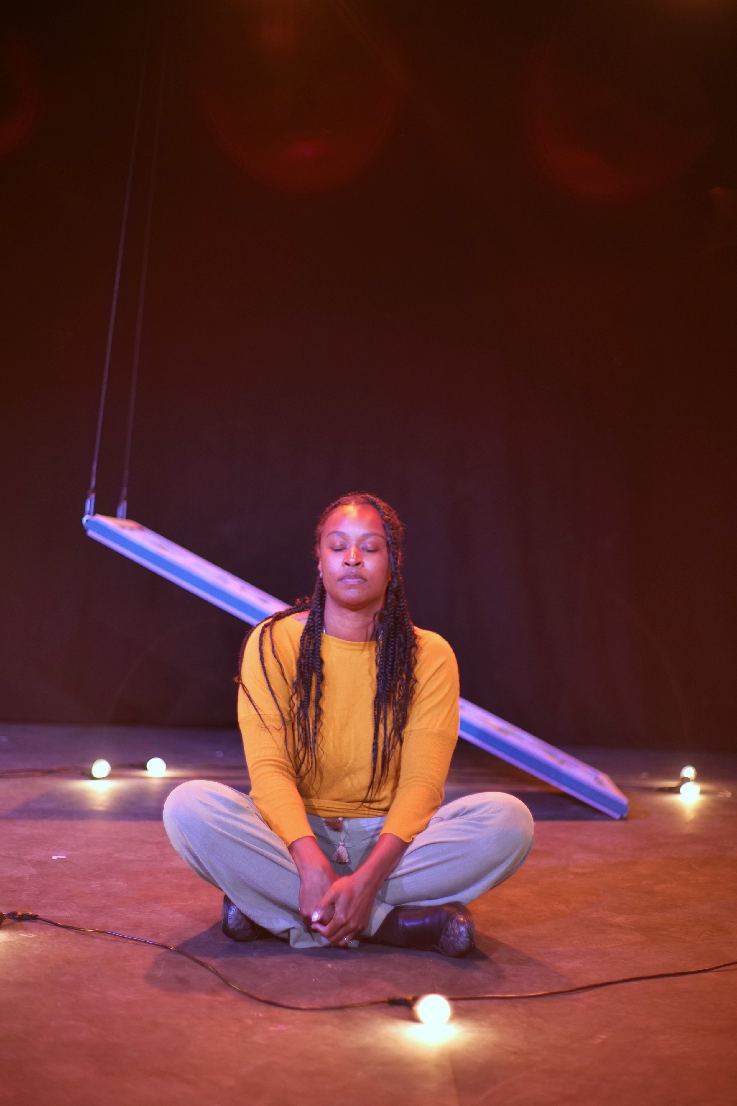  Photo credit: Ryd Cook  A black woman sits cross legged with her eyes closed. A string of light bulbs encircle the ground around her. Behind her, a door held up by strings has fallen onto the floor on one side. 